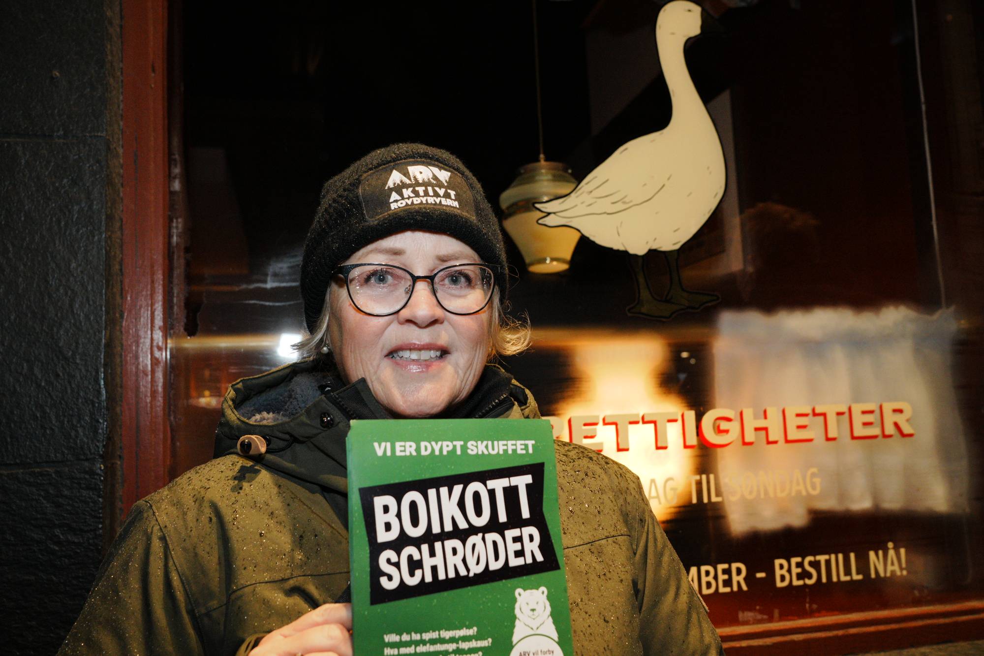 Kvinne holder plakat med teksten Boikott Schrøder foran en restaurant med gåseillustrasjon i vinduet. (Bildebeskrivelsen er laget av en KI-tjeneste)