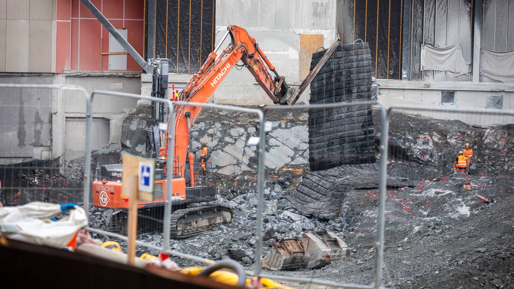 Anleggsmaskin løfter en sprengningsmatte på byggeplass med arbeidere i bakgrunnen. (Bildebeskrivelsen er laget av en KI-tjeneste)