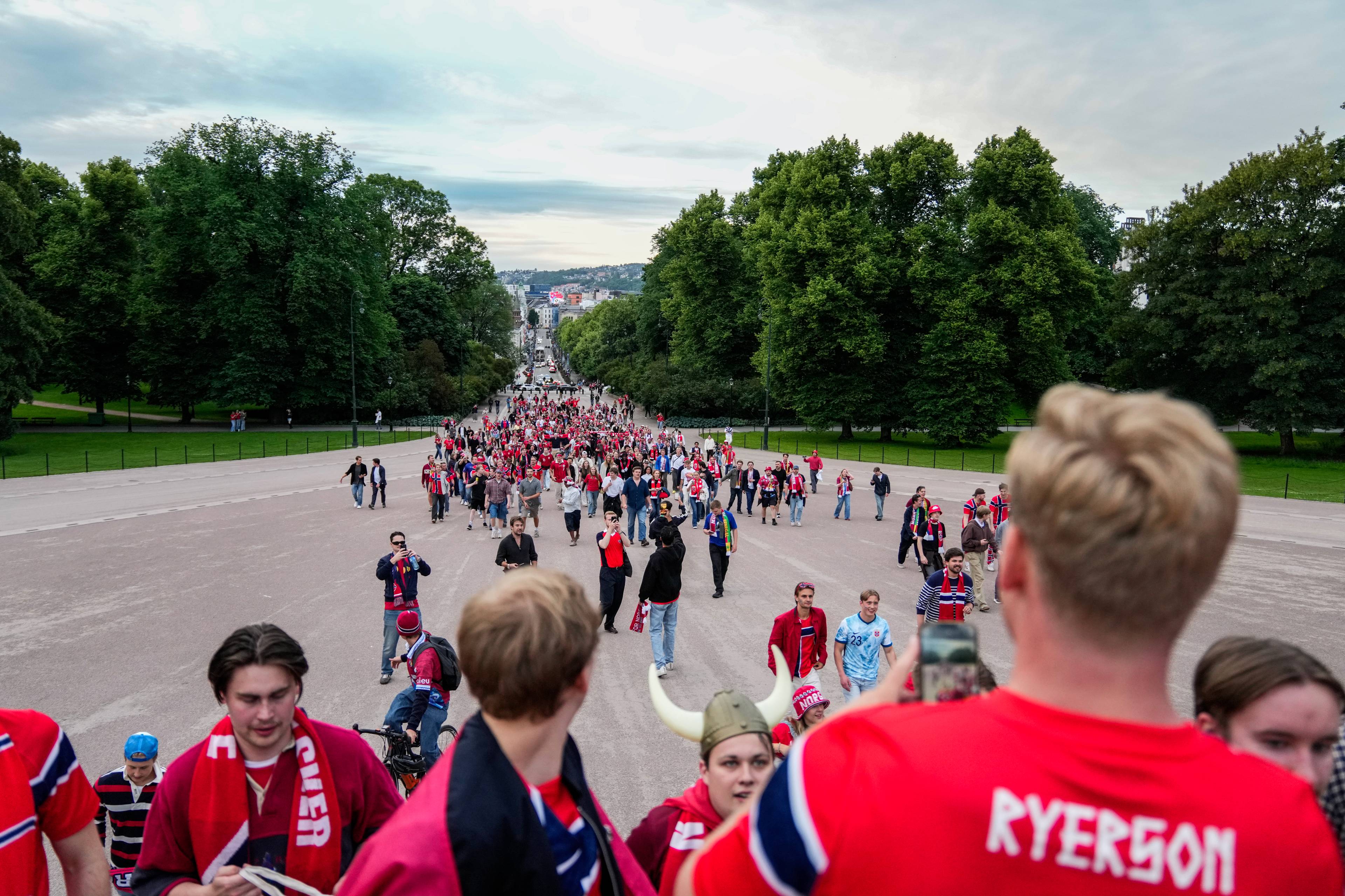 Folk i røde og hvite supporterklær går i prosesjon opp en bred grusvei med trær på sidene. En gruppe mennesker, de fleste med norske flagg og skjerf, går forbi. En mann i forgrunnen tar bilde av folkemengden med telefonen. Landskapet åpner seg opp med utsikt over en by i det fjerne.