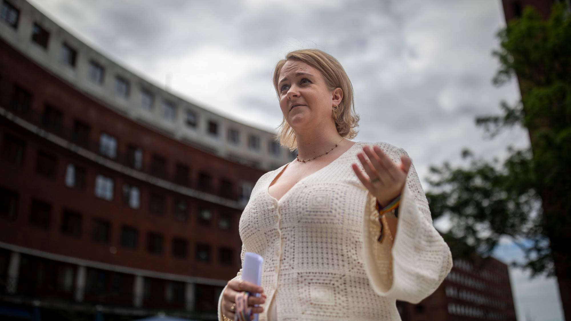 En kvinne i hvit heklet kjole holder en lilla gjenstand i venstre hånd. Hun gestikulerer med høyre hånd mens hun snakker utendørs. Bak henne sees en mørk mursteinsbygning med mange vinduer. (Bildebeskrivelsen er laget av en KI-tjeneste)