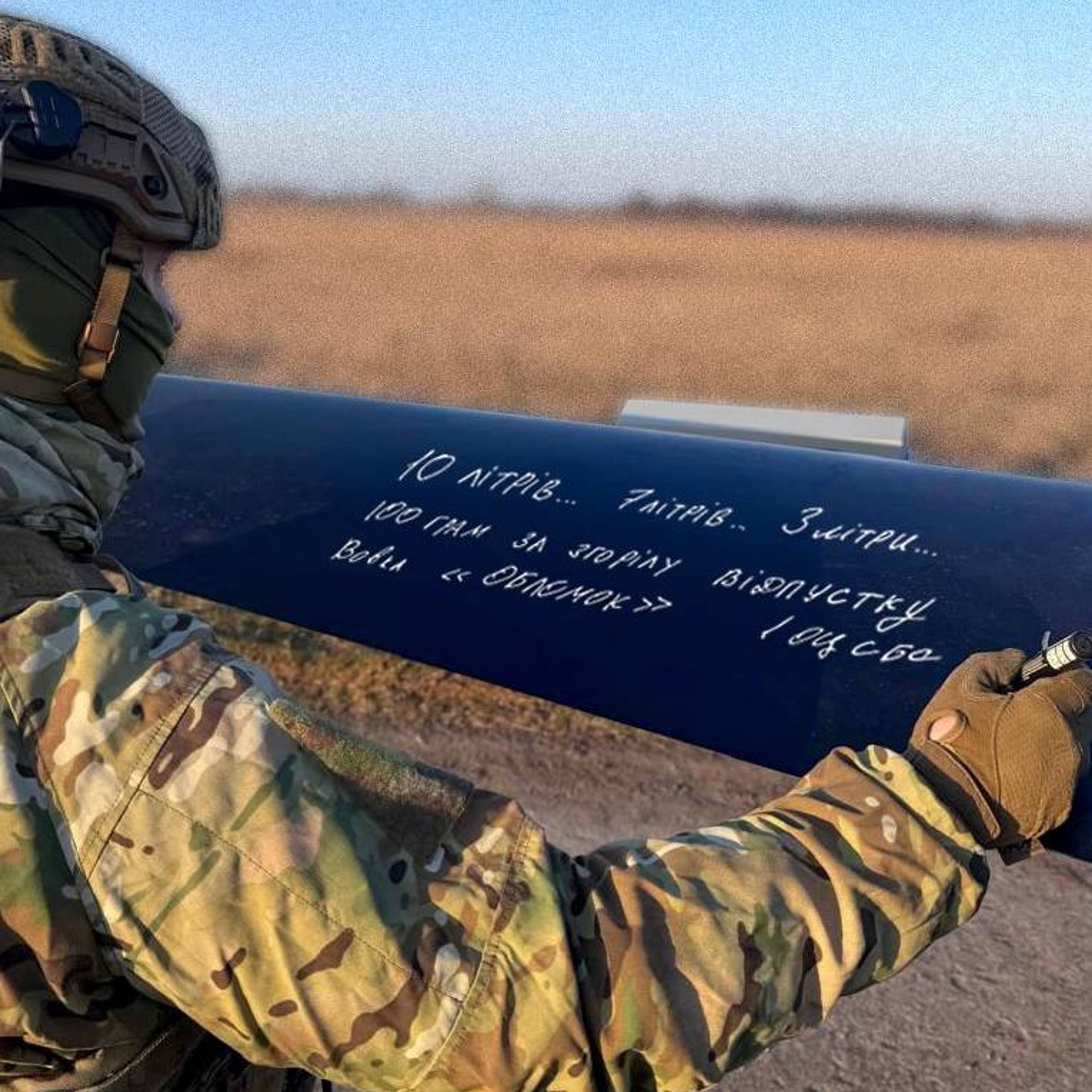 En soldat skriver en melding på en rakett. Soldaten er ikledd kamuflasjeuniform og hjelm. Raketten ligger på bakken i et åpent landskap. (Bildebeskrivelsen er laget av en KI-tjeneste)