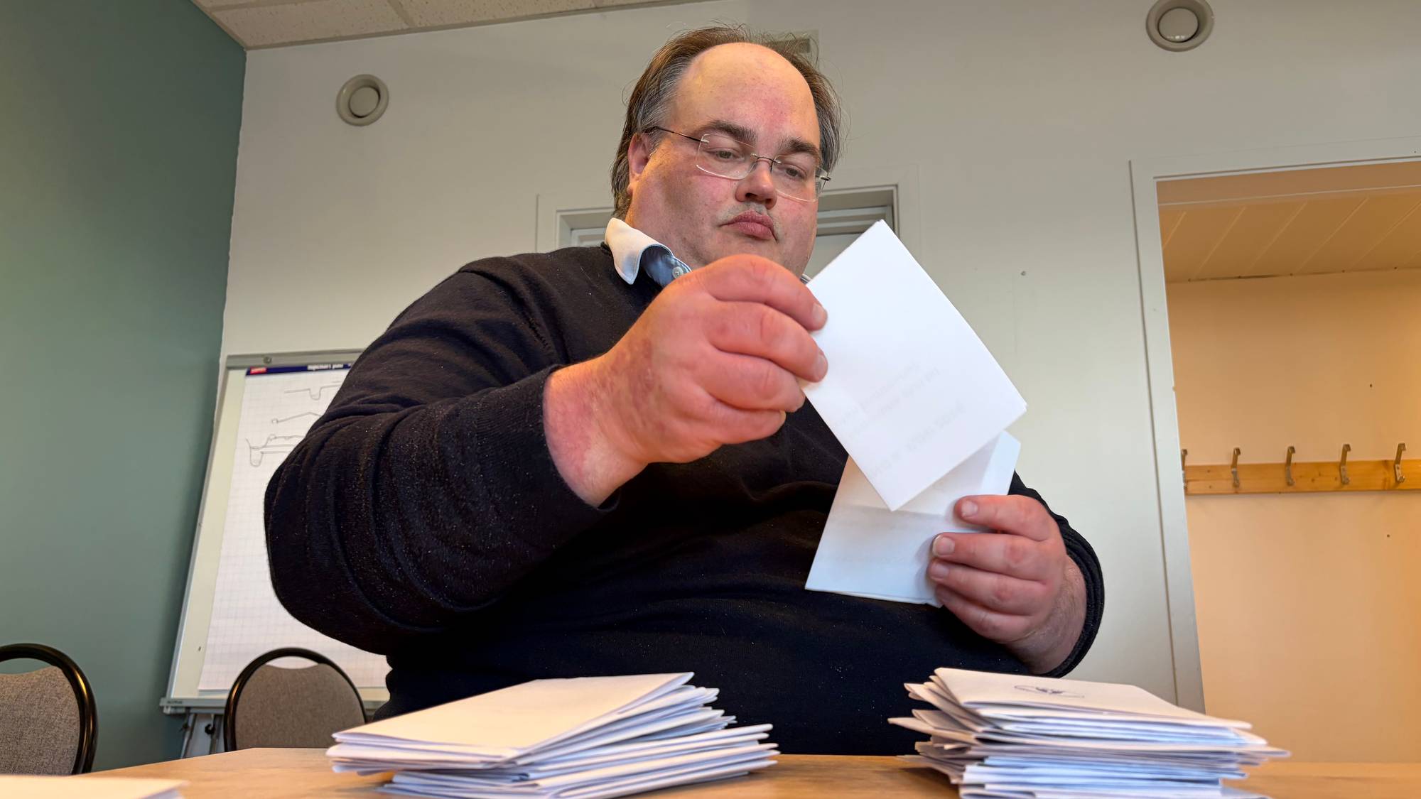 En mann med briller sorterer papirer i et møterom. Han holder et hvitt ark i den ene hånden og et hvitt konvolutt i den andre. Rundt ham ligger flere konvolutter stablet på et trebord. Bak mannen er en whiteboardtavle. (Bildebeskrivelsen er laget av en KI-tjeneste)