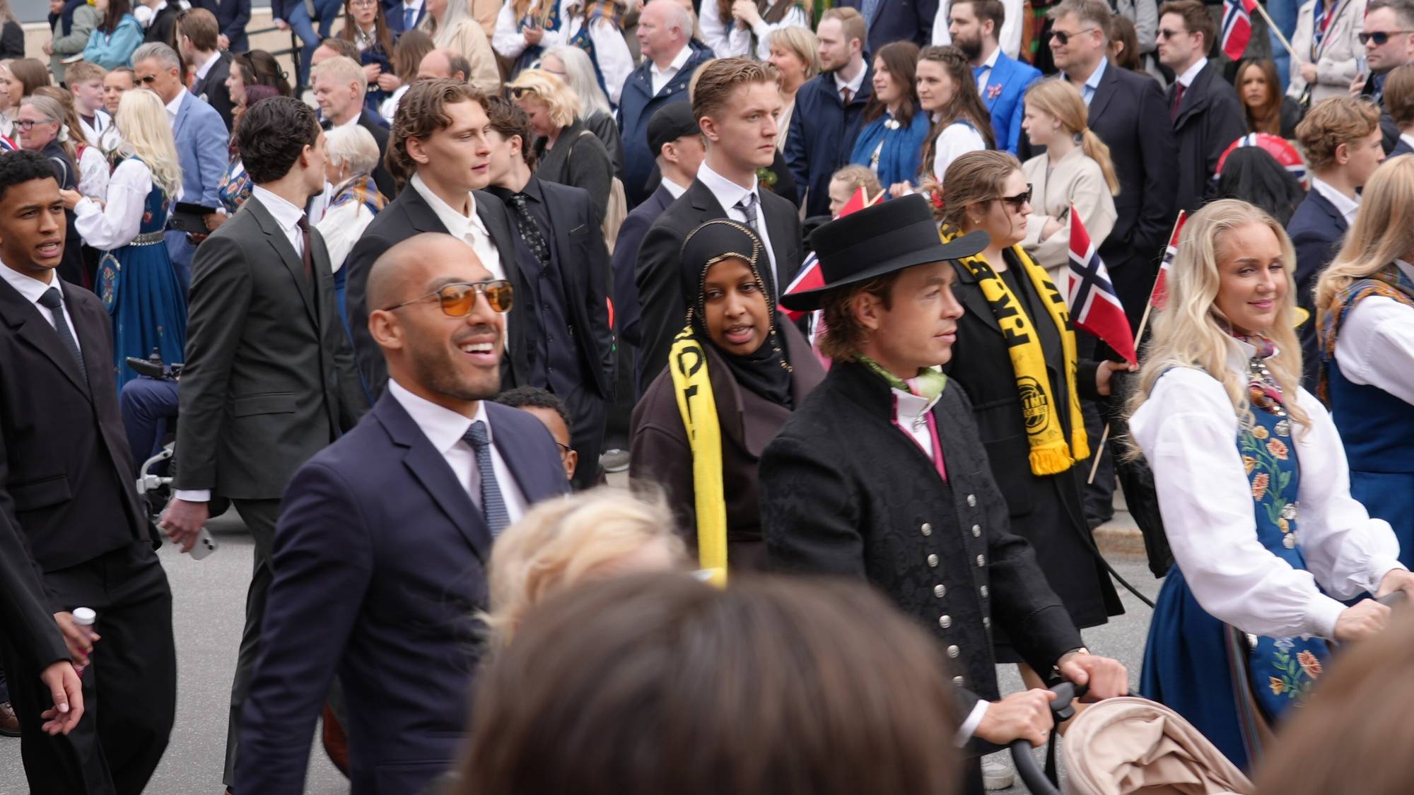 En gruppe mennesker går langs en gate, mange kledd i bunad og dress. Noen holder norske flagg. I forgrunnen står menn i dress og kvinner i bunad. Bildet viser en folkemengde i et utendørs arrangement. (Bildebeskrivelsen er laget av en KI-tjeneste)