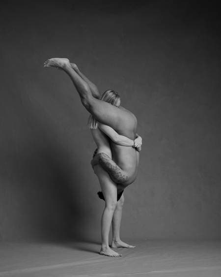 En blond kvinne står oppreist og bærer en mann med mørkt hår og tatoveringer opp ned foran sin kropp. Han har ansiktet mot skrittet hennes. Begge er nakne, men ingen kjønnsorganer er synlige.