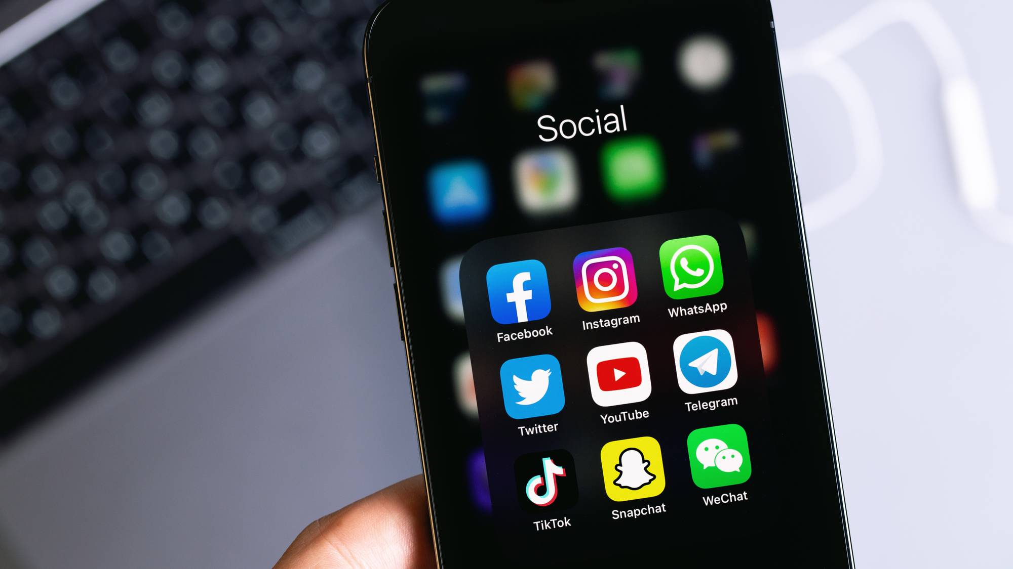 En mobiltelefon viser ikonene for sosiale medieapper. Ikonene inkluderer Facebook, Instagram, WhatsApp, Twitter, YouTube, Telegram, TikTok, Snapchat og WeChat. Telefonen holdes av en hånd, og en tastatur er synlig i bakgrunnen. Bakgrunnen er uskarpt hvit. (Bildebeskrivelsen er laget av en KI-tjeneste)