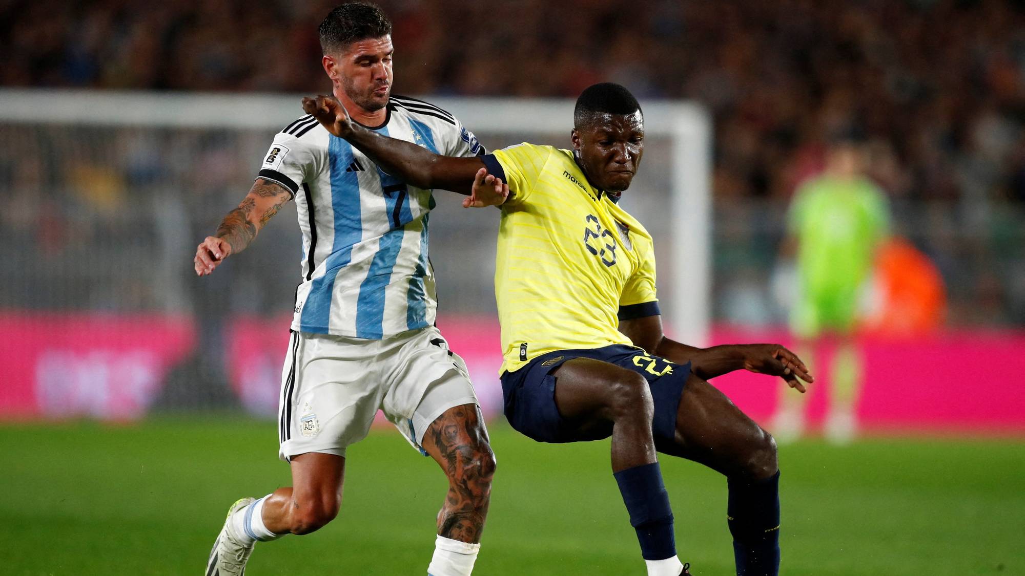 To fotballspillere i aksjon på en gressbane. Den ene spilleren er iført Argentinas hvite og lyseblå drakt og den andre spilleren er iført Ecuadors gule drakt. Bakgrunnen er utydelig med publikum og en del av et mål. (Bildebeskrivelsen er laget av en KI-tjeneste)