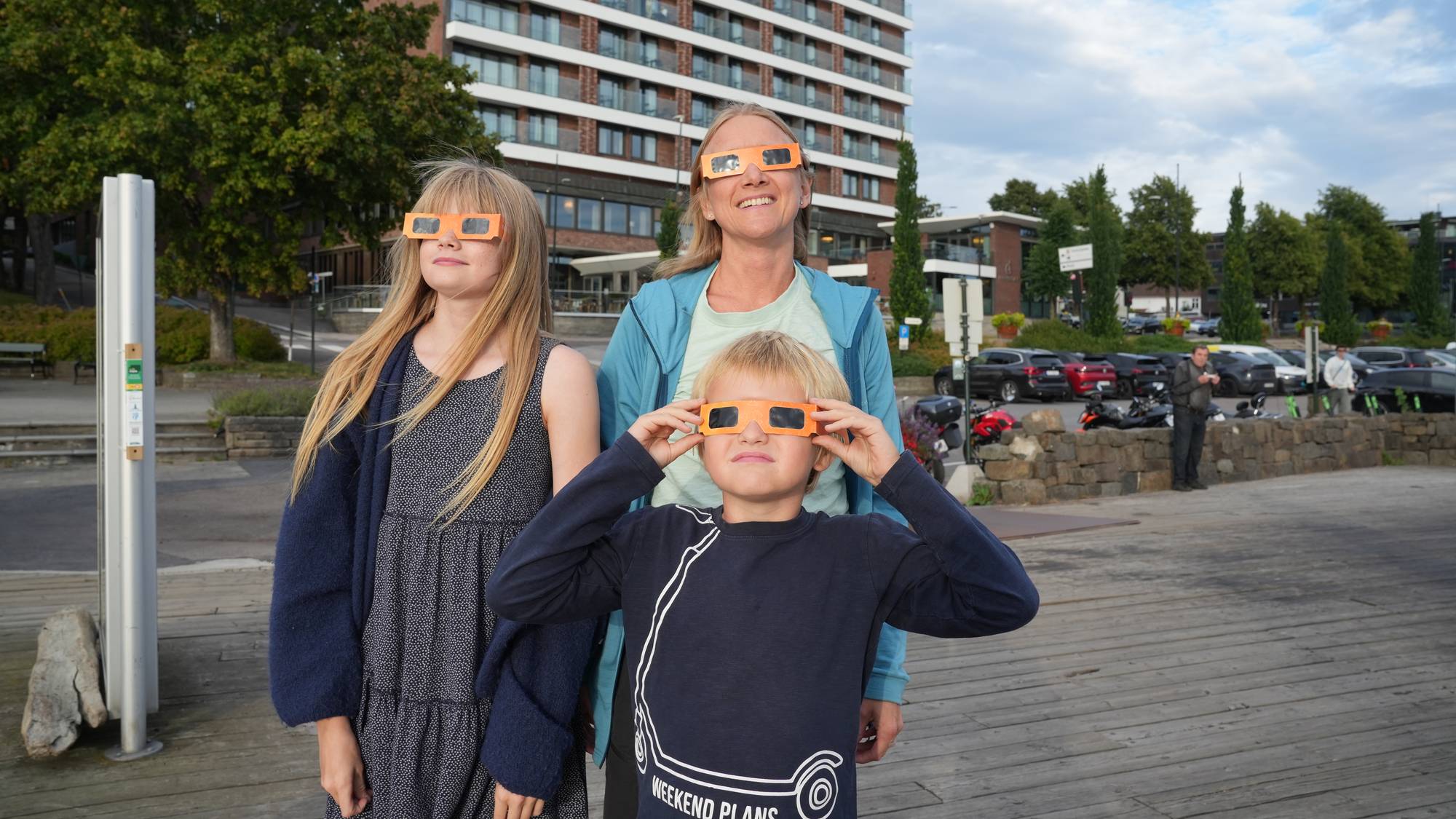 Kvinne og to barn ser på solformørkelse med beskyttelsesbriller ved et hotell. (Bildebeskrivelsen er laget av en KI-tjeneste)