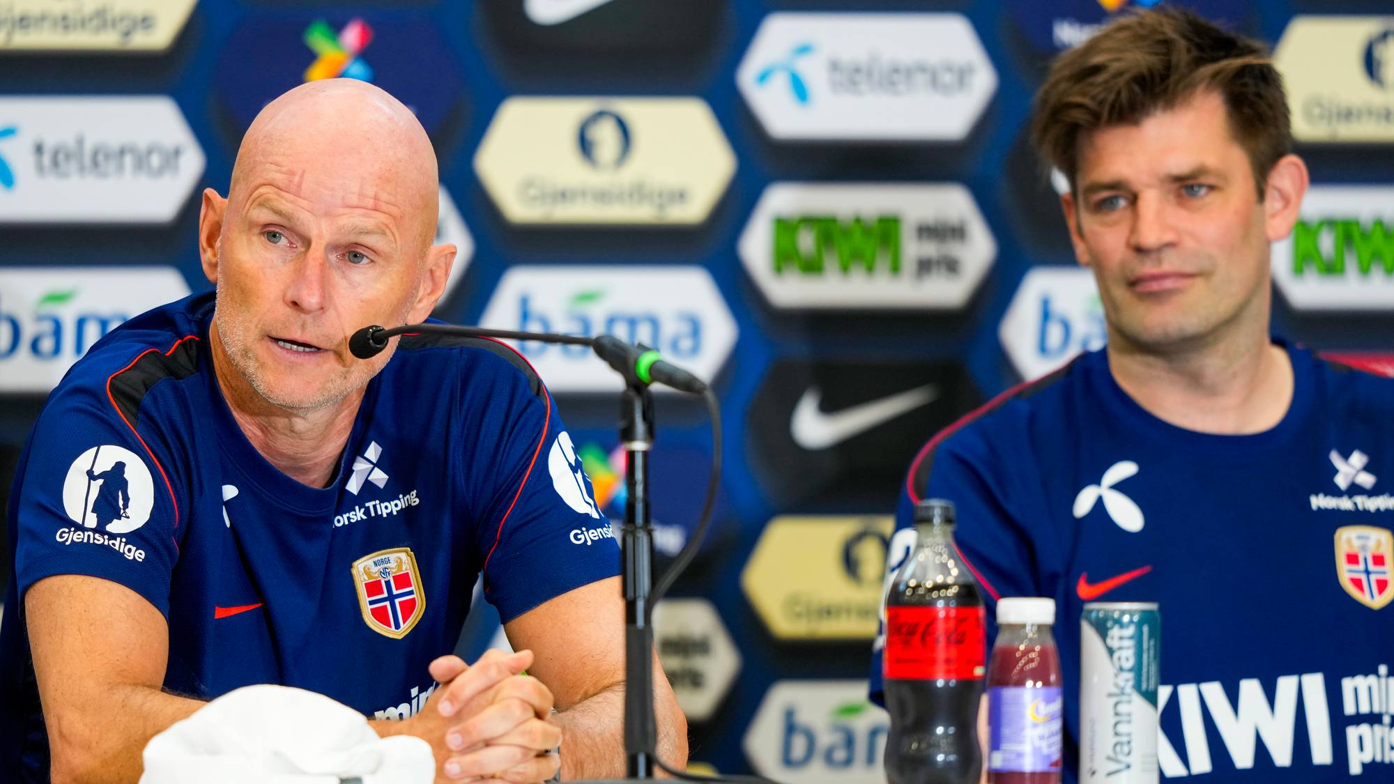 To menn sitter bak et bord med drikkevarer. Den ene mannen, med skallet hode, snakker inn i en mikrofon. Begge mennene har på seg mørkeblå t-skjorter med norske flagg og sponsorlogoer. Bak dem er en bakvegg med flere logoer. (Bildebeskrivelsen er laget av en KI-tjeneste)