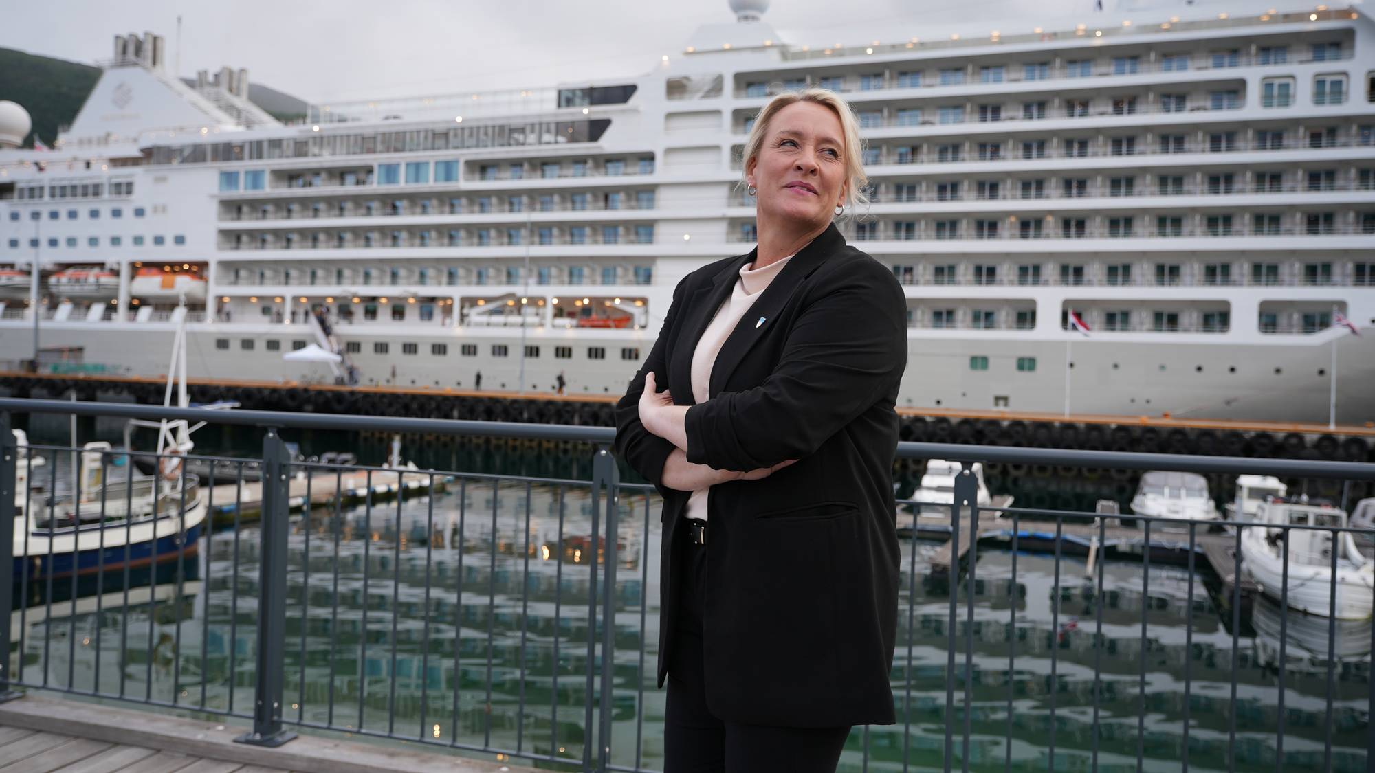 En kvinne i svart dressjakke står med armene i kors. Bak henne ligger et stort cruiseskip til kai, og i forgrunnen skimtes flere mindre båter. Det er en grå og overskyet dag. (Bildebeskrivelsen er laget av en KI-tjeneste)
