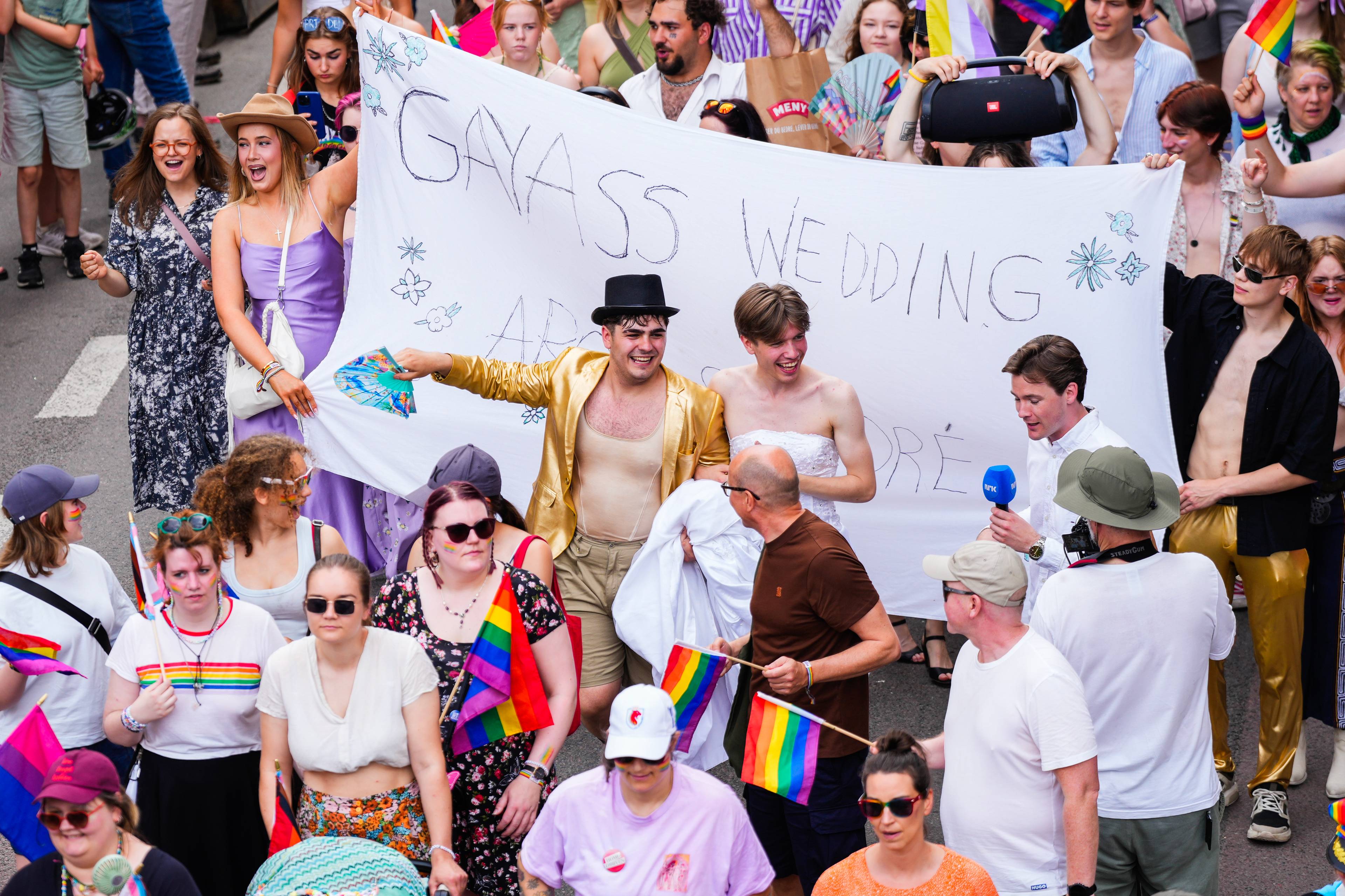 Folkemengde i Pride-parade bærer et skilt med teksten Gayass wedding. Flere holder regnbueflagg. (Bildebeskrivelsen er laget av en KI-tjeneste)