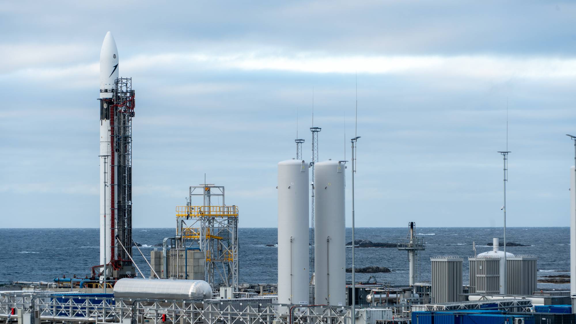 Rakett står klar på en utskytningsrampe ved havet med industrielle tanker i forgrunnen. (Bildebeskrivelsen er laget av en KI-tjeneste)