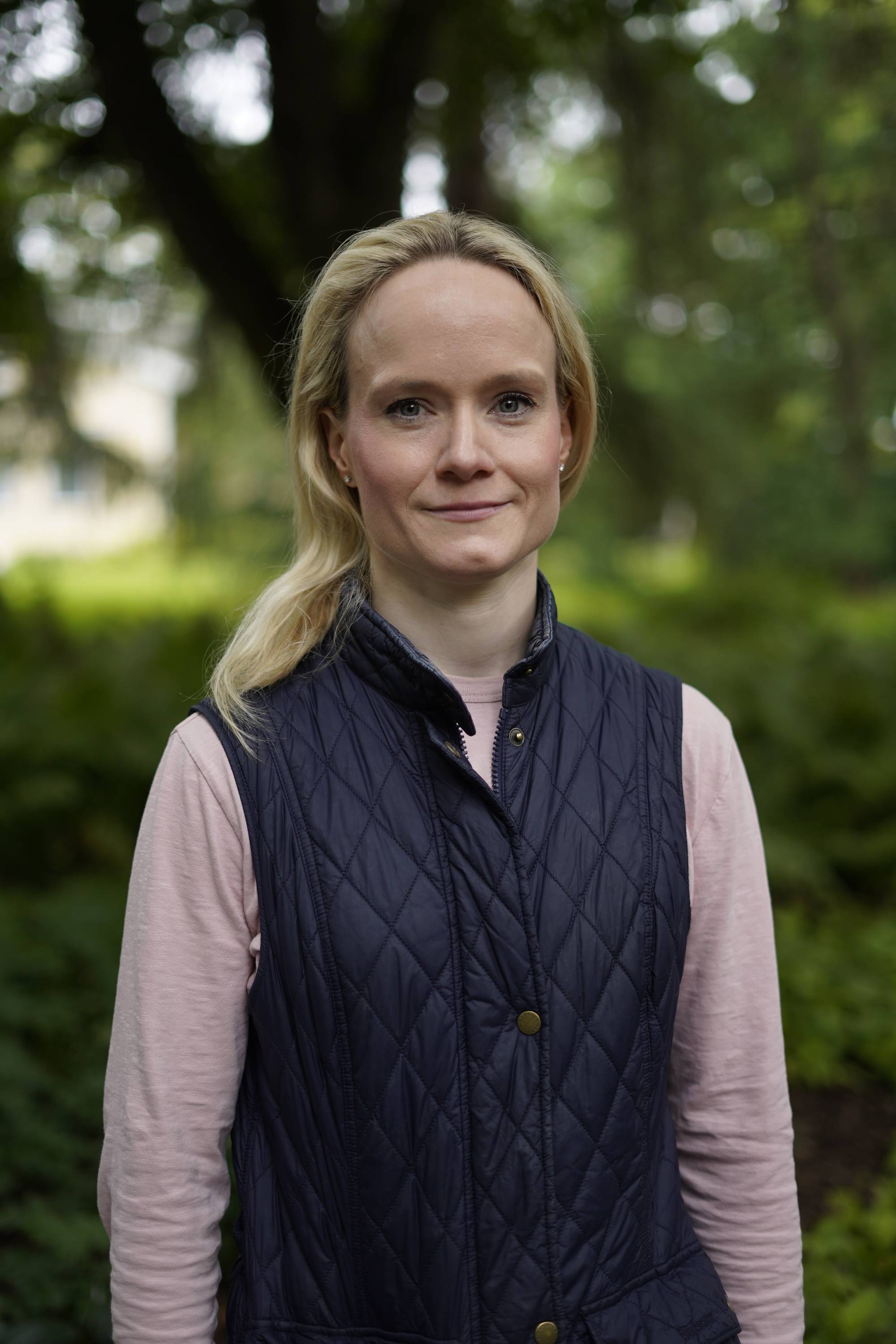 En kvine med lys rosa genser under en blå vest, ser rett i kamera- Hun står i en park md grønne trær i bakgrunnen. 