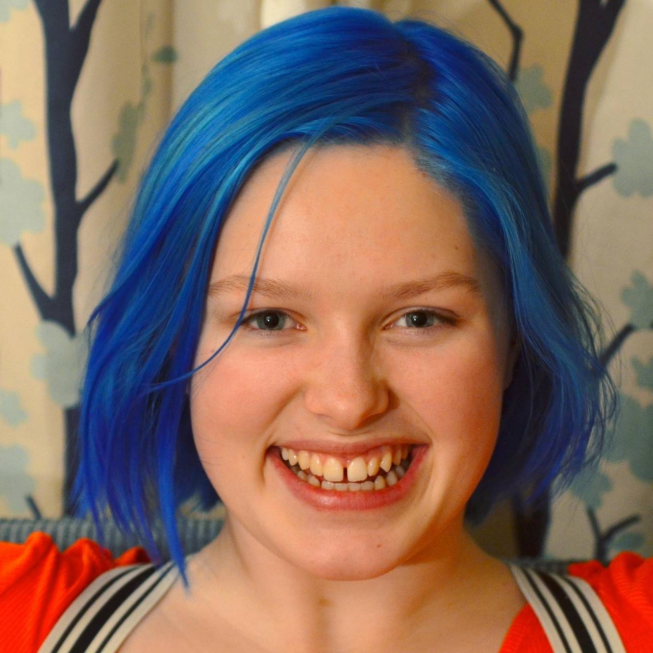 På bildet har Aurora Leigh Kobernus blått, halvlangt hår. Hun smiler. I bakgrunnen er det lyse gardiner med trær på. 
