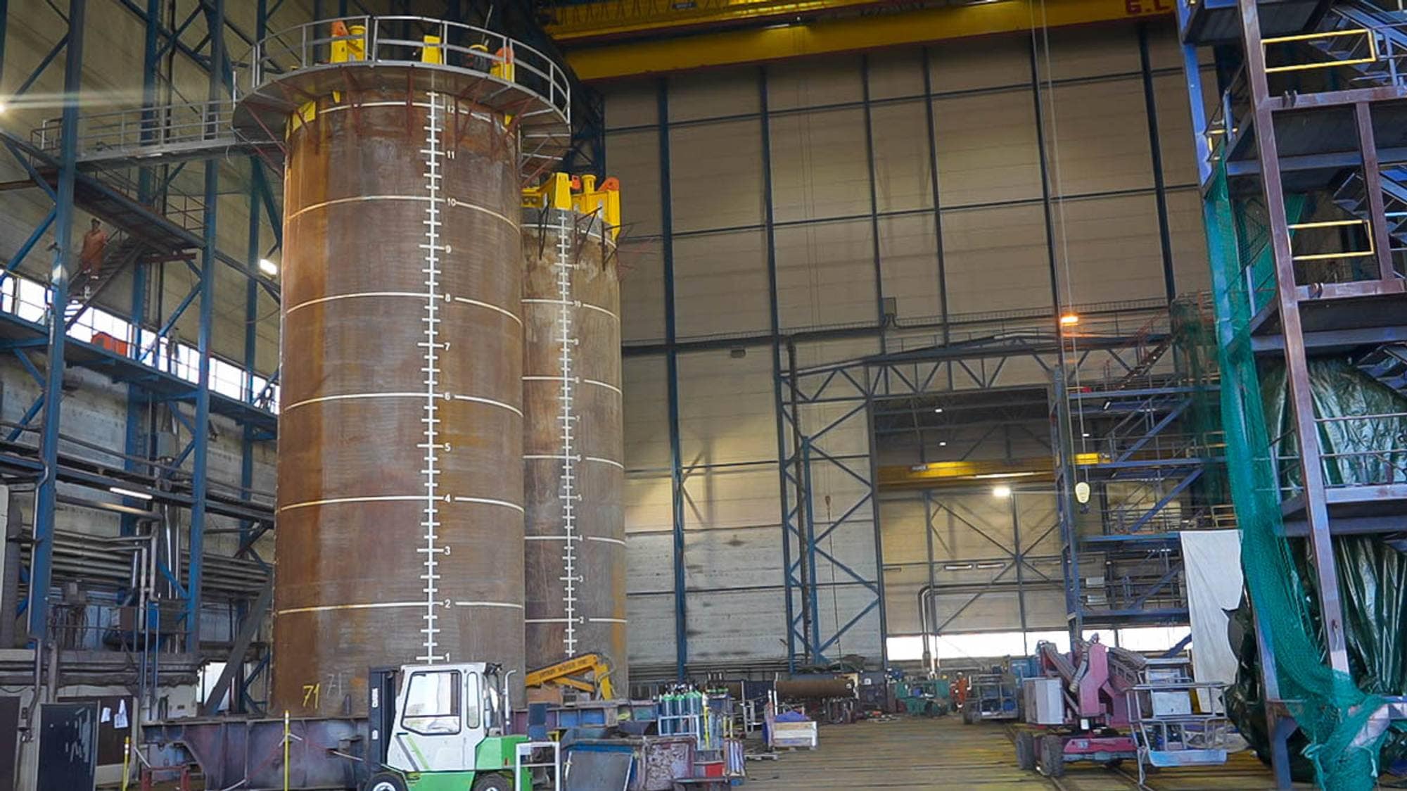 To store sylindriske metalltanker merket med måleskala i en stor verkstedhall. (Bildebeskrivelsen er laget av en KI-tjeneste)