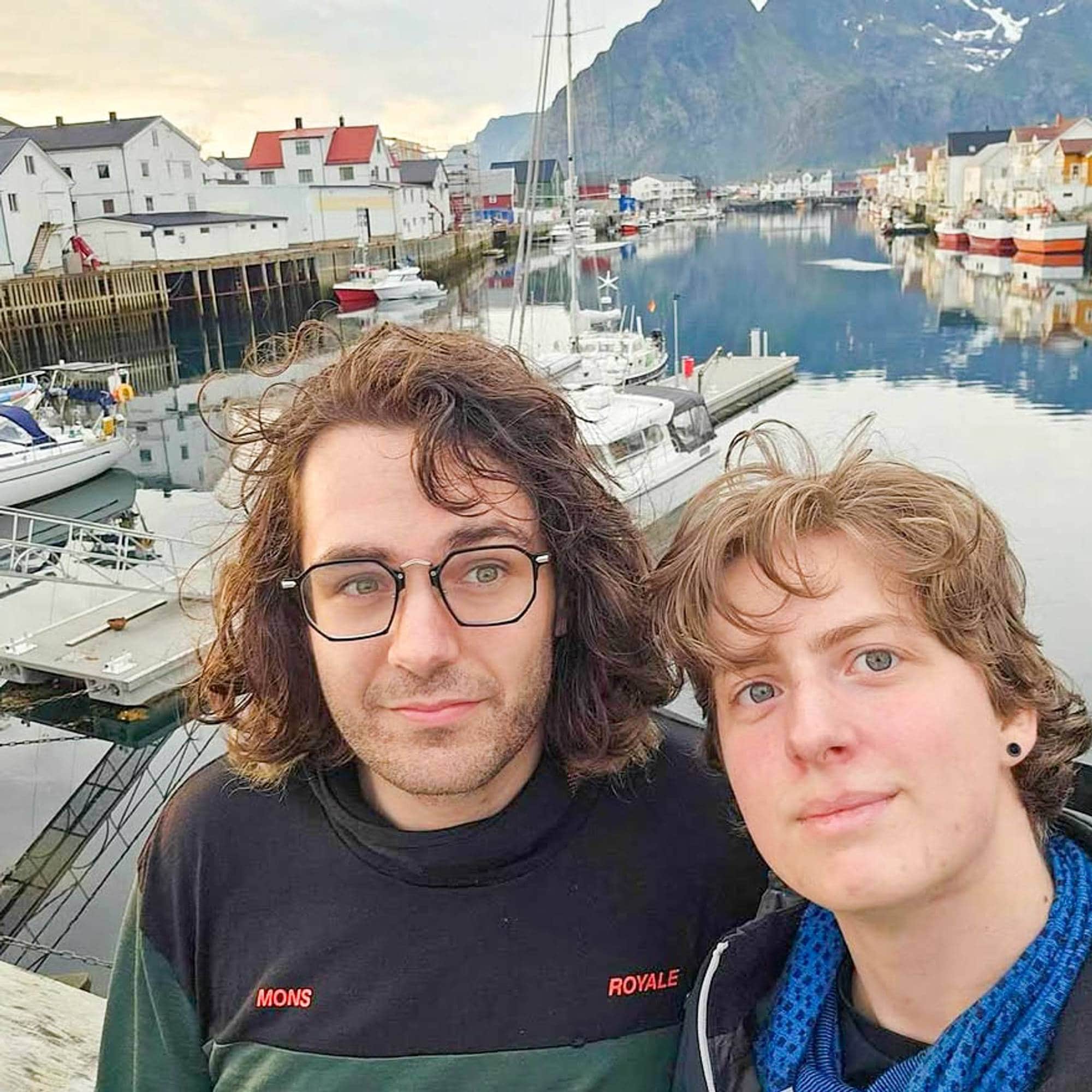 To persongar, ein med halvlangt hår og brillar og ein med øredobb, ser mot kamera. I bakgrunn ser ein fjord og fjell ved Henningsvær med fleire båtar og kvite hus som spegler seg i vatnet.