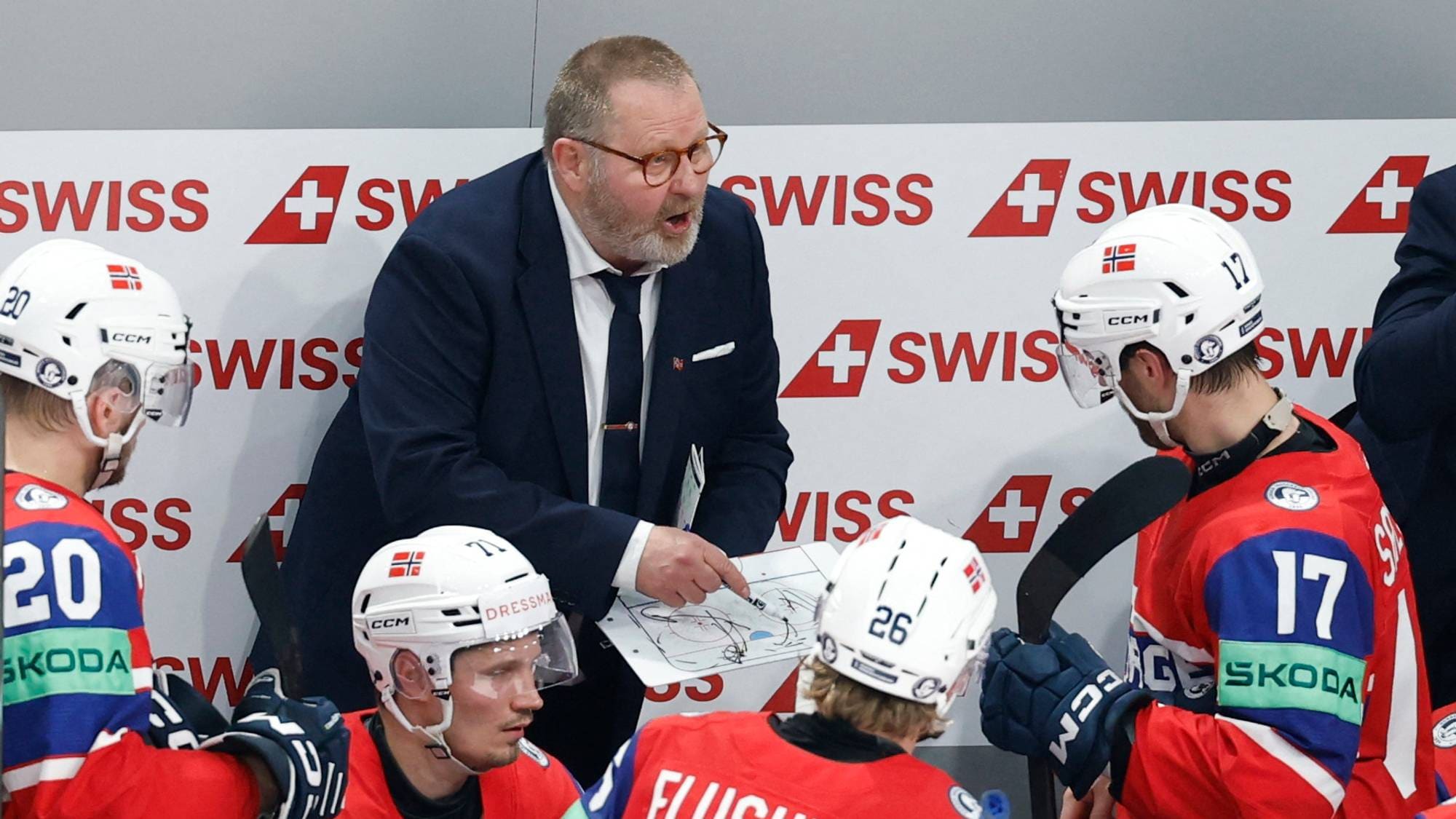 En ishockeytrener gir instruksjoner til spillere. Treneren holder opp et taktikkbrett med tegninger. Spillerne er iført landslagsdrakter med nummer på ryggen. Bak dem er en reklamevegg med Swiss-logo. (Bildebeskrivelsen er laget av en KI-tjeneste)