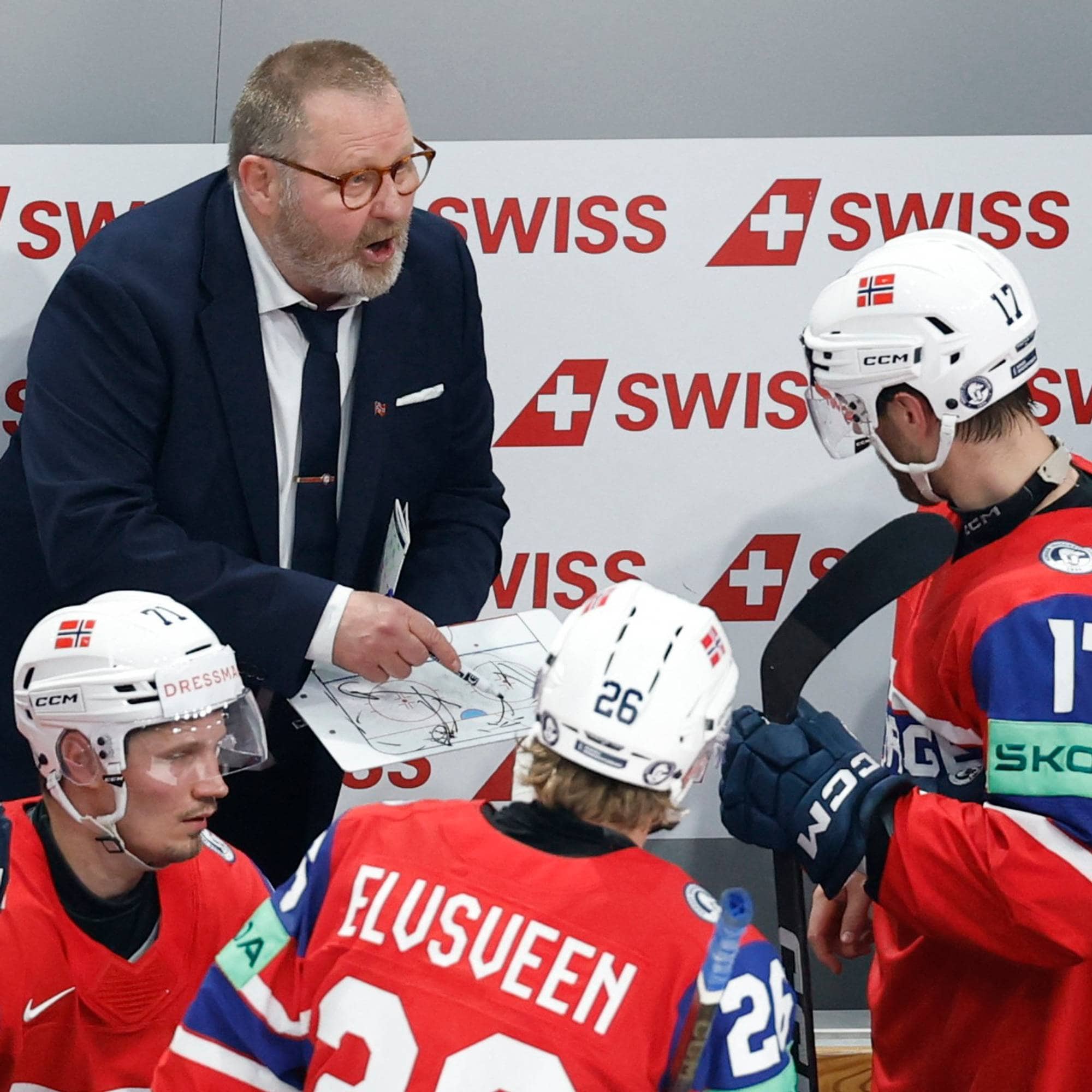 En ishockeytrener gir instruksjoner til spillere. Treneren holder opp et taktikkbrett med tegninger. Spillerne er iført landslagsdrakter med nummer på ryggen. Bak dem er en reklamevegg med Swiss-logo. (Bildebeskrivelsen er laget av en KI-tjeneste)