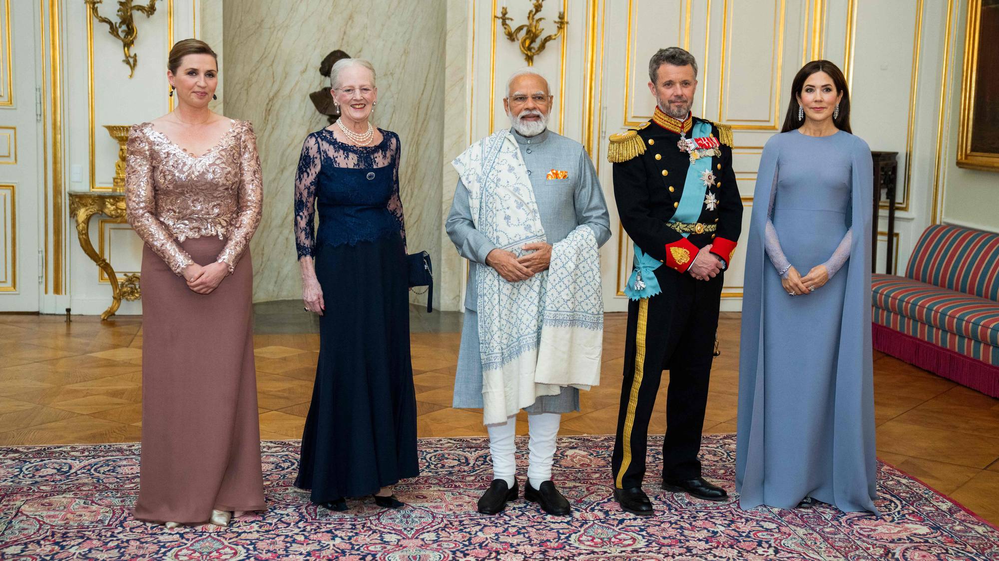 Statsminister Mette Frederiksen, dronning Margrethe, Indias statsminister Narendra Modi, kronprins Frederik og kronprinsesse Mary står sammen. De er kledd i formelle antrekk. Bildet er tatt innendørs i et rom med gullfargede detaljer og en stor, mønstret teppegulv. (Bildebeskrivelsen er laget av en KI-tjeneste)