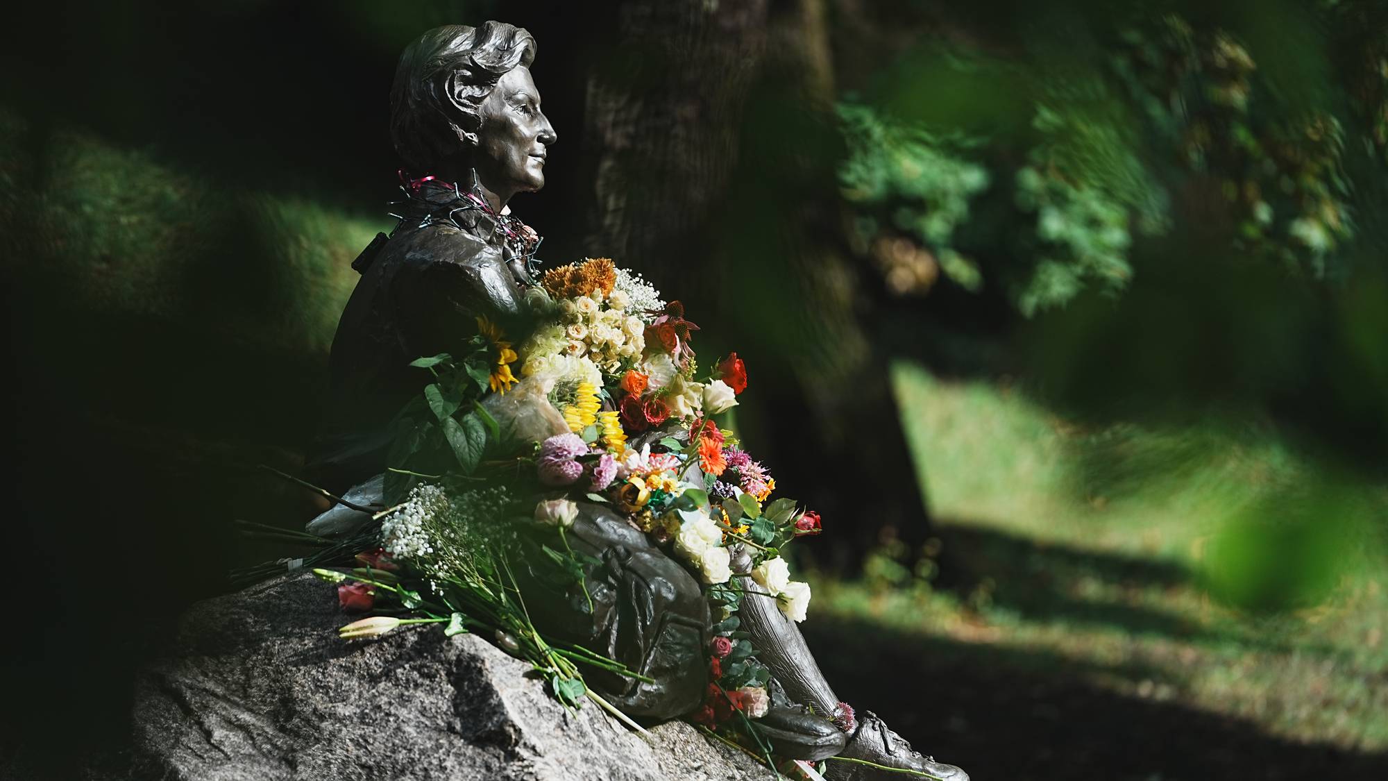 Bronzestatue av kvinne med blomsterbuketter plassert i en park. (Bildebeskrivelsen er laget av en KI-tjeneste)