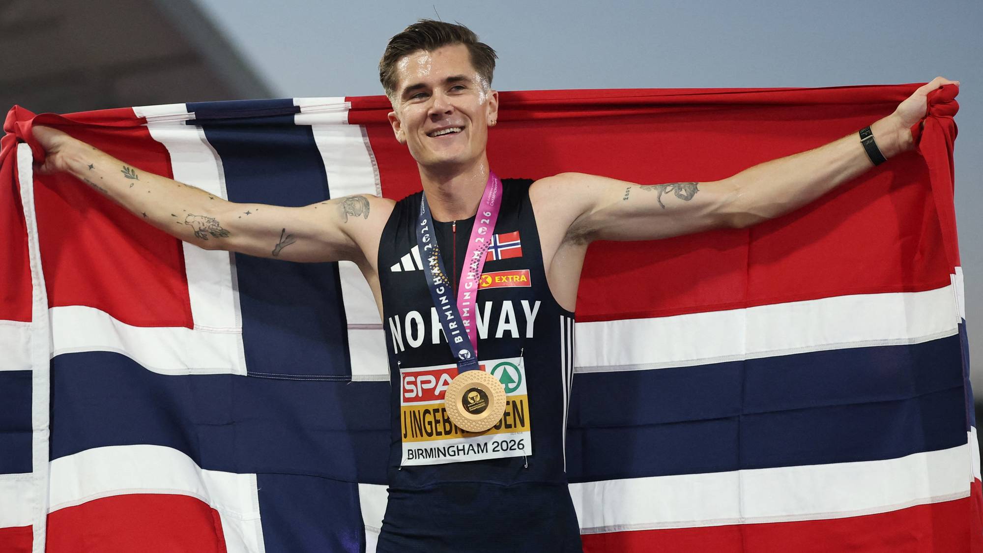 Jakob Ingebrigtsen smiler og holder opp et norsk flagg etter gulløp på en friidrettsbane. (Bildebeskrivelsen er laget av en KI-tjeneste)