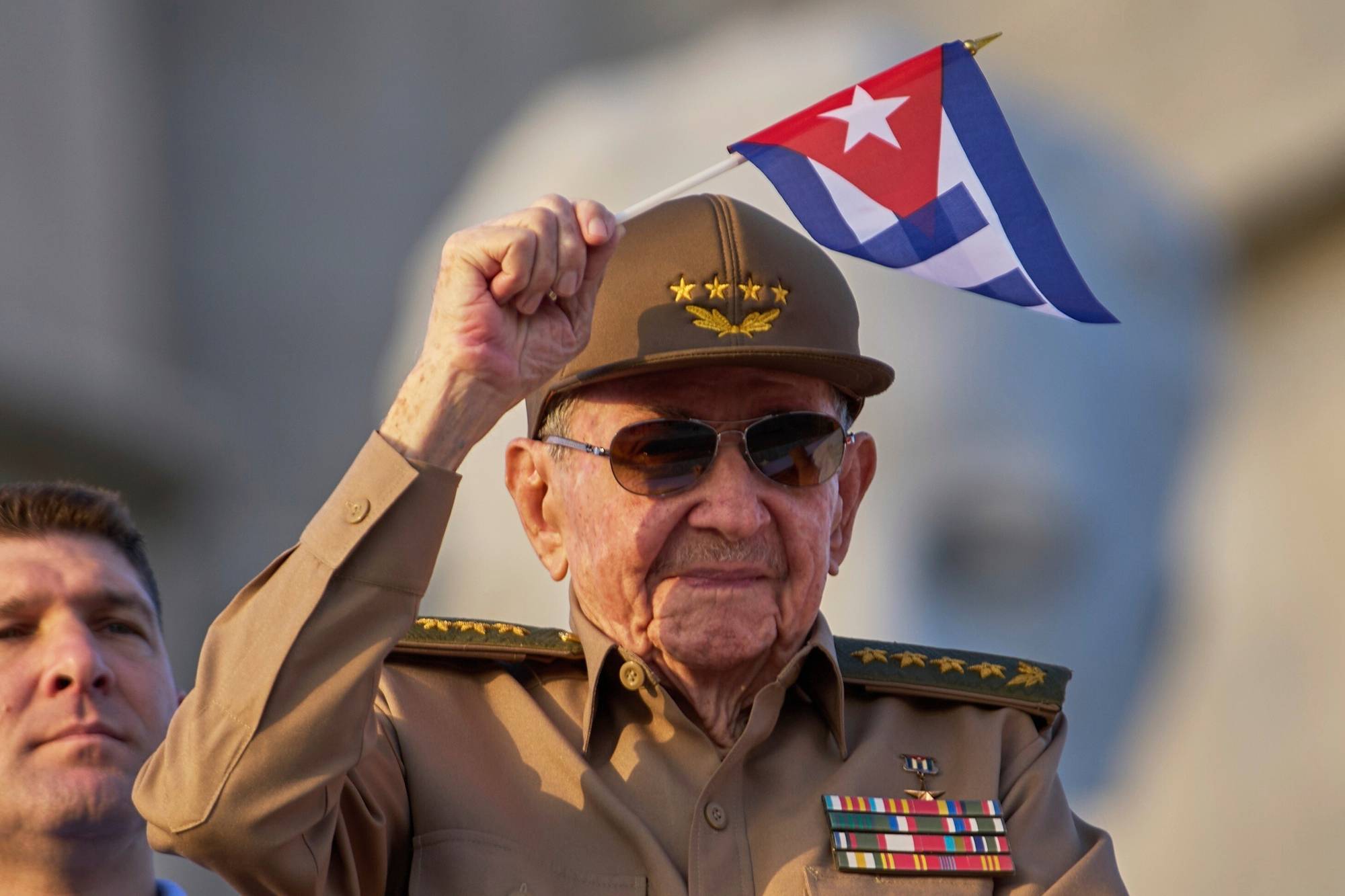 En eldre mann i militæruniform holder et cubansk flagg. Han har på seg en brun uniform med medaljer og en hatt. En annen mann sees til venstre i bildet. (Bildebeskrivelsen er laget av en KI-tjeneste)
