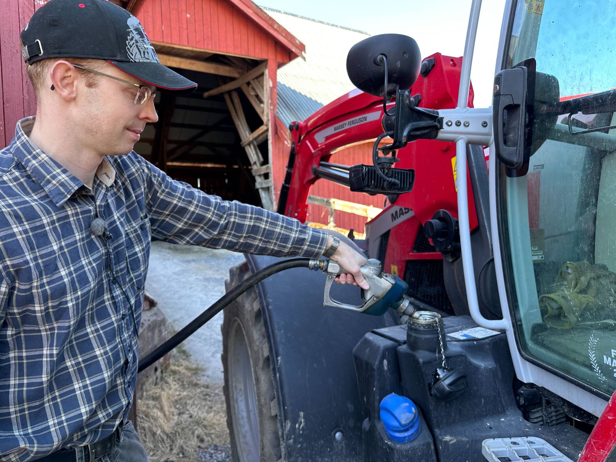 En bonde fyller på bensin på en traktor. I bakgrunnen kan man se en rød låve.
