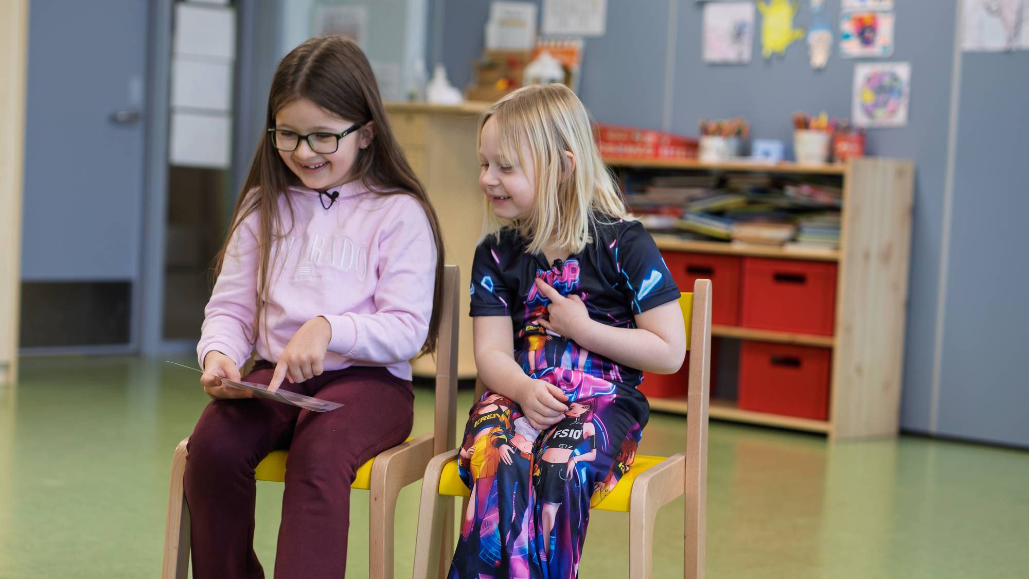 To jenter ser på et bilde. Valentina har mørkt hår og holder bilder i hånden. Marie har blondt hår og ser på bildet. De smiler begge to. De sitter på stoler midt i et klasserom. I bakgrunnen ser man en blå vegg med tegninger på veggen. 