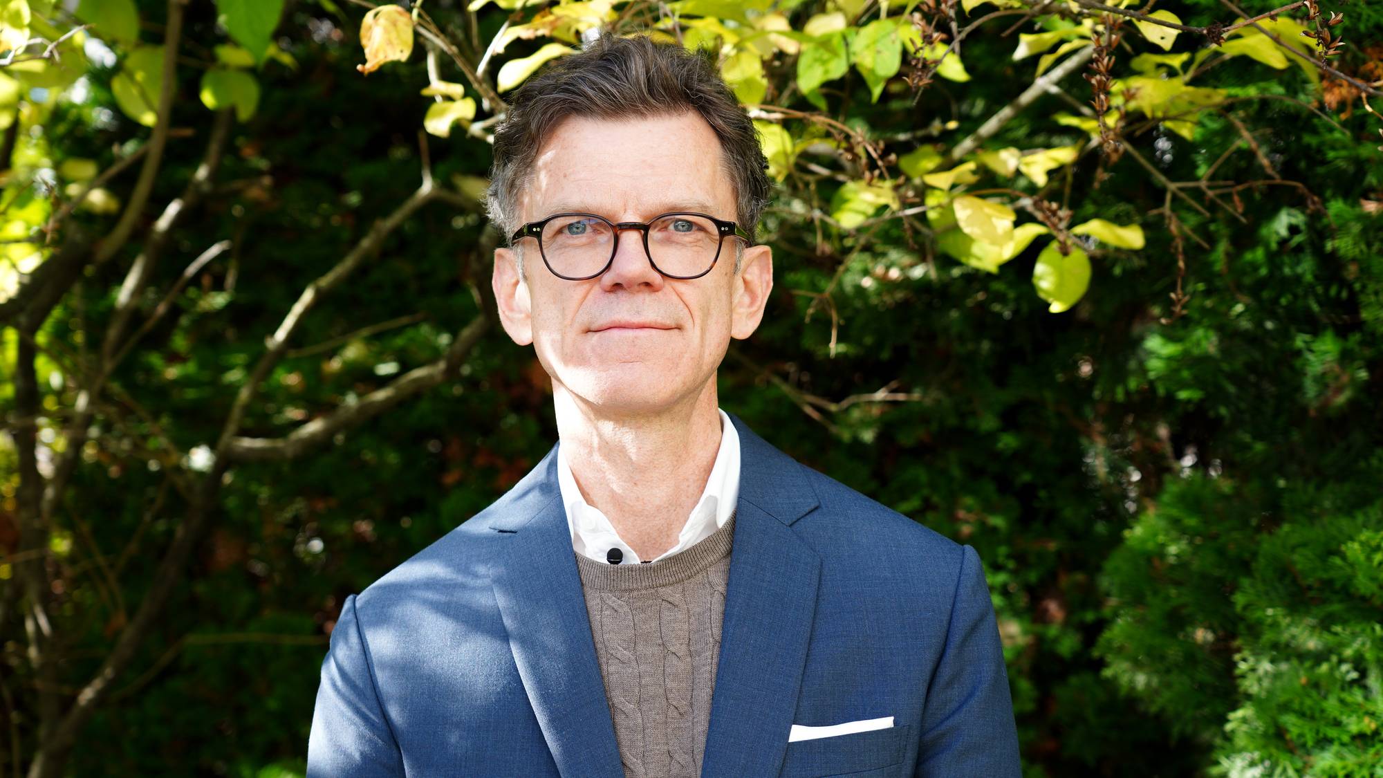 Petter-Børre Furberg som stiller opp til portrett utenfor en grønn plantevegg, kledd i blå dressjakke og en hvit skjorte med en genser over den. 