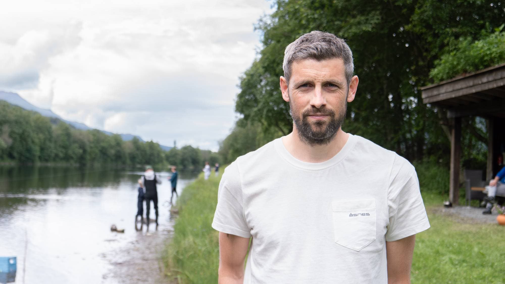 En mann står i forgrunnen ved en elv, der andre personer fisker i bakgrunnen. (Bildebeskrivelsen er laget av en KI-tjeneste)