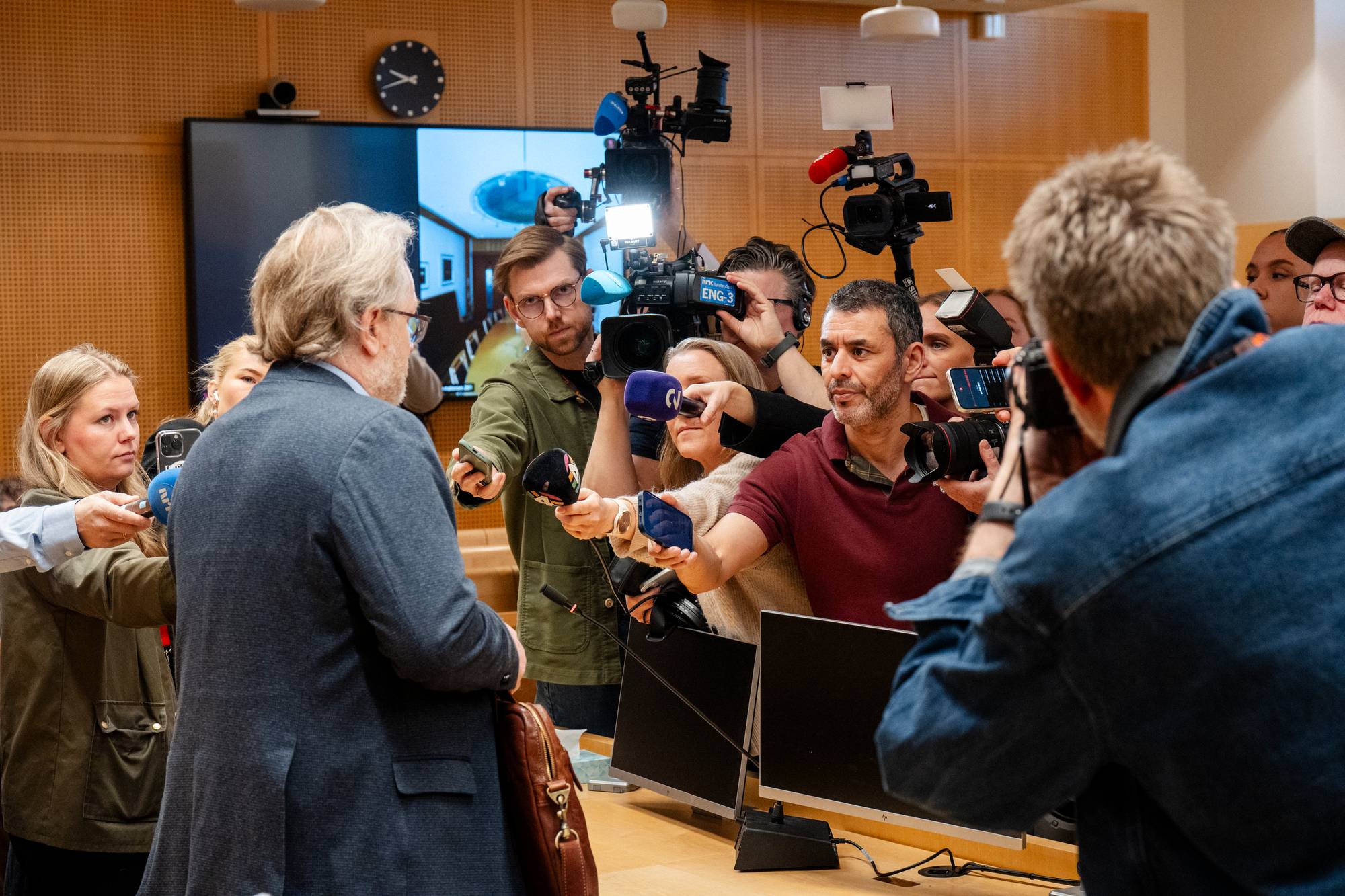 En mann med dress blir intervjuet av journalister. Mange mikrofoner er rettet mot ham. Bak journalister står mange kameraer. (Bildebeskrivelsen er laget av en KI-tjeneste)