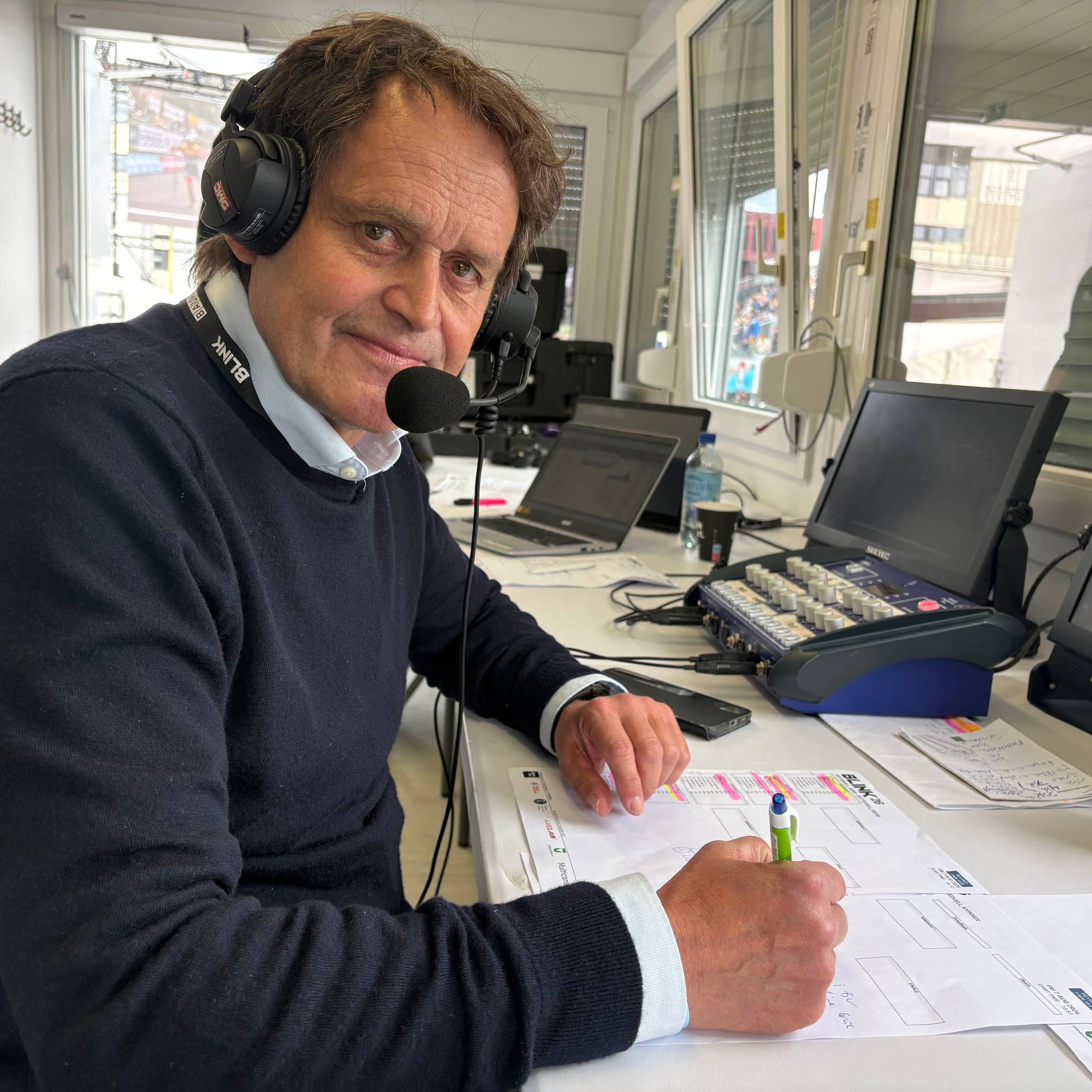 En mann med blå genser ser inn i kameraet. På hodet har han et headset og i hånda en penn. På bordet foran han ligger det diverse papir med skrift. 