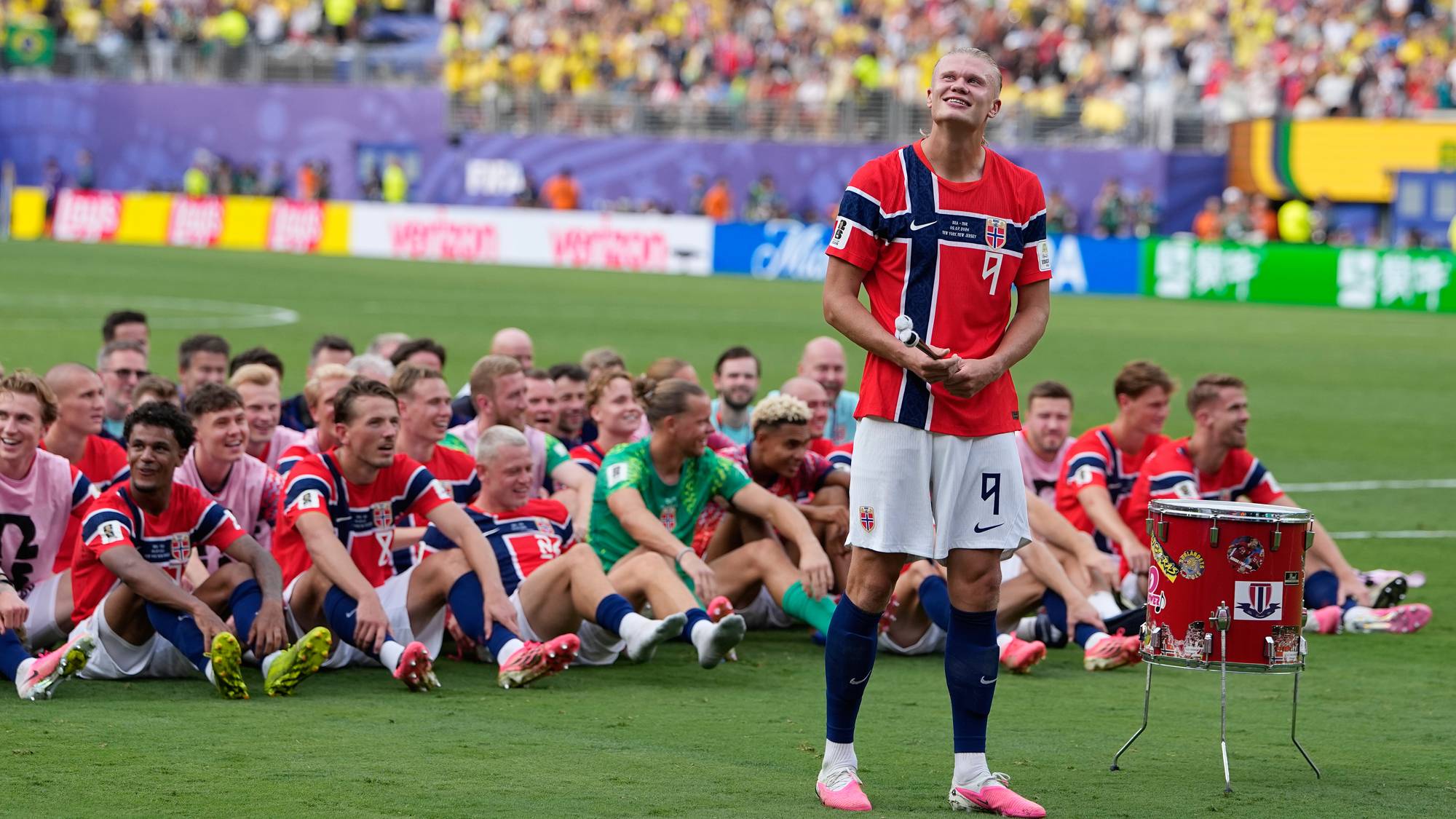 Erling Braut Haaland står på gresset med trommestikker foran sitt sittende norske fotballag. (Bildebeskrivelsen er laget av en KI-tjeneste)