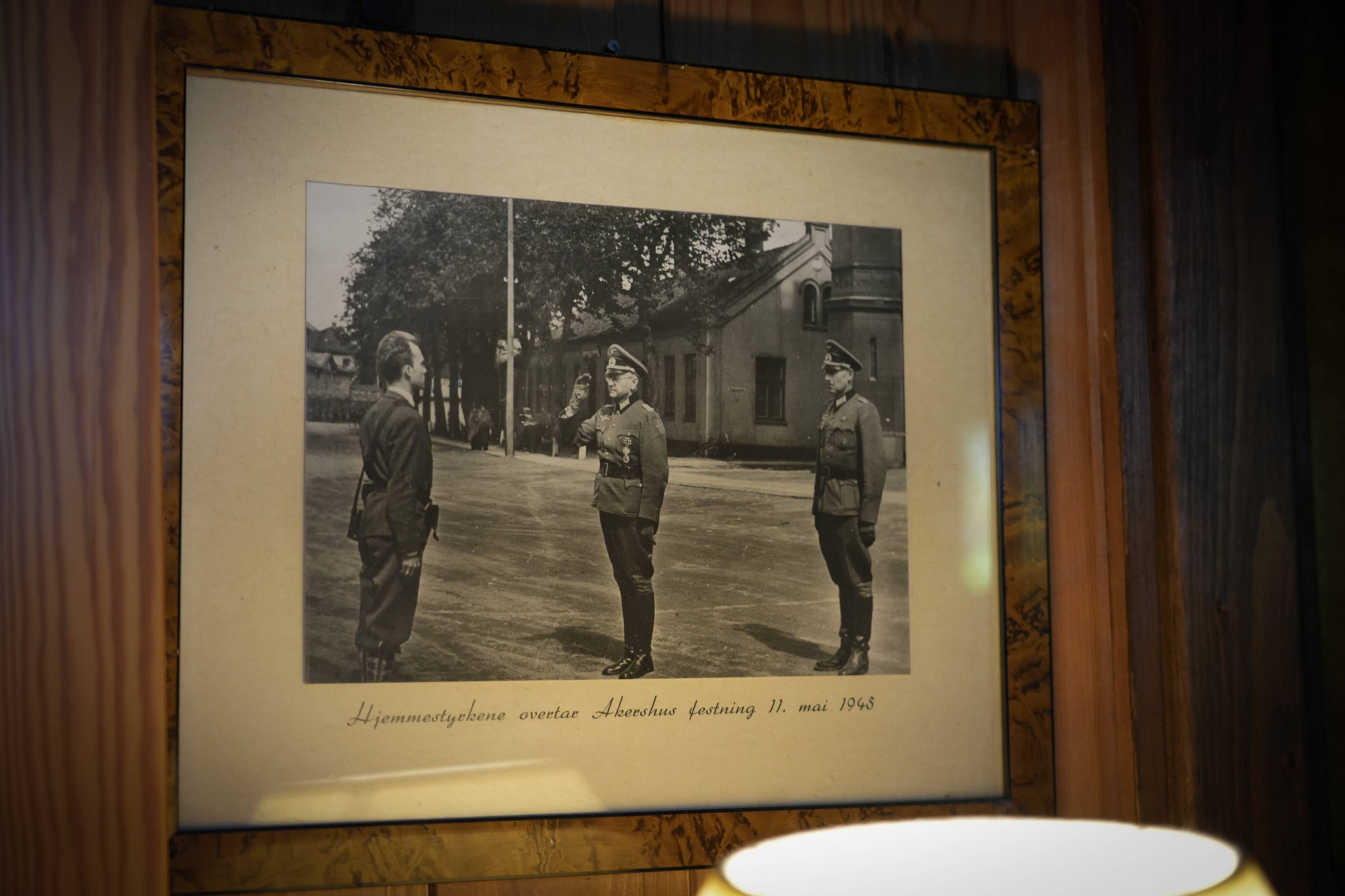 Et svart-hvitt bilde viser tre menn i militæruniform. En mann i midten hilser. I bakgrunnen ses bygninger og trær. Teksten under bildet lyder "Hjemmestyrkene overtar Akershus festning 11. mai 1945". (Bildebeskrivelsen er laget av en KI-tjeneste)