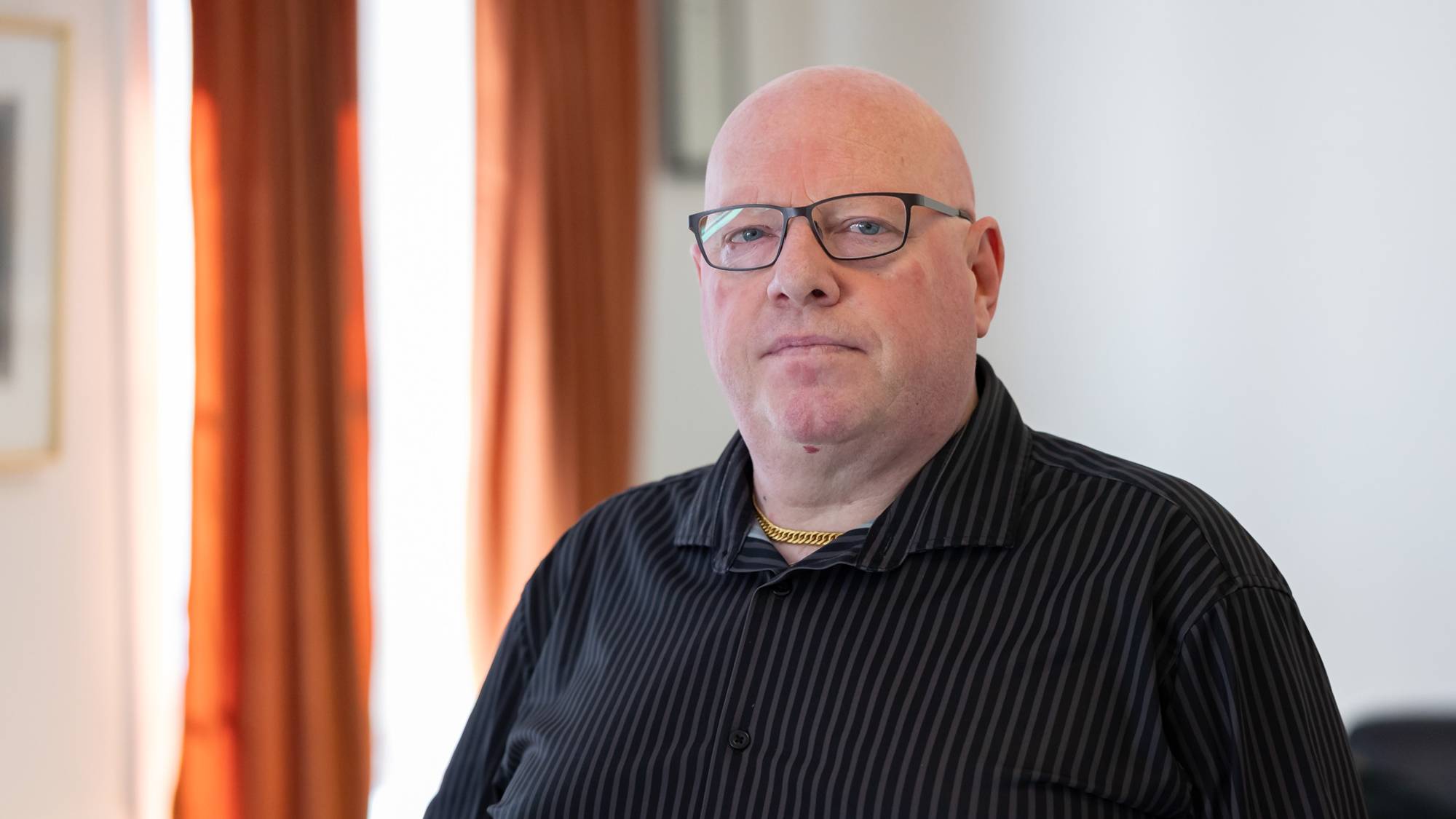 En mann iført svart skjorte med hvite striper og en brille med mørk innfatning står og kikker mot fotografen. Han har ikke hår på hodet. Han står i et rom som ligner en stue. Vi kan se noen oransje gardiner i bakgrunnen og deler av et bilde på veggen