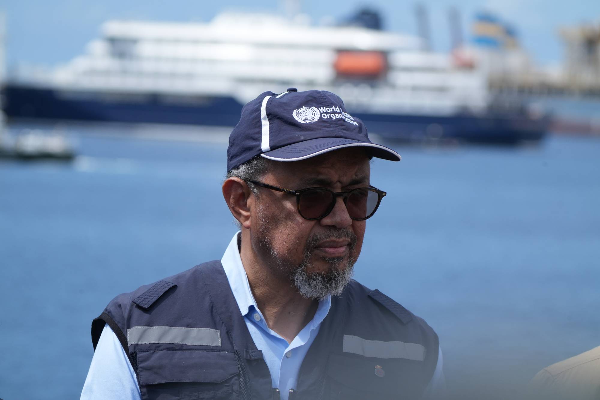 Mann med caps og solbriller. Han har på seg en blå vest over en lyseblå skjorte. I bakgrunnen sees et stort skip i en havn. Mannen ser ned til høyre. (Bildebeskrivelsen er laget av en KI-tjeneste)