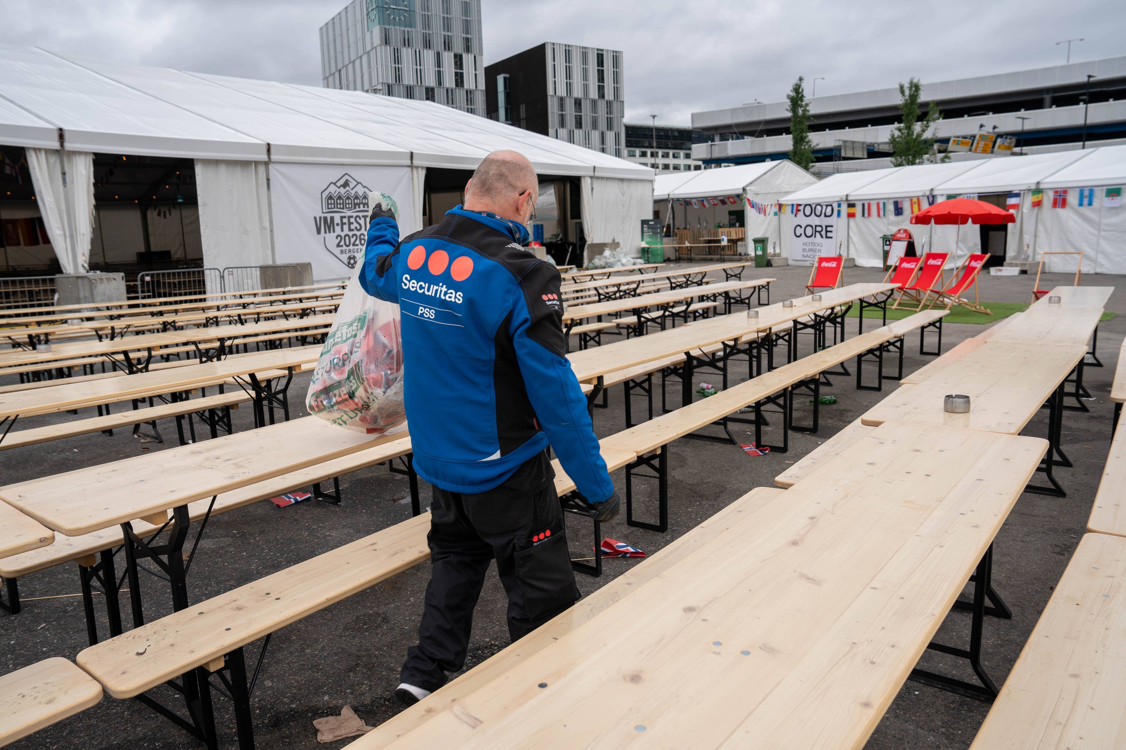 Ein Securitas-tilsett ber ein stor plastsekk med søppel. Han går forbi mange lange trebord og benker utanfor ein teltbygning. På ein av teltveggene står det «VM-FEST 2026 BERGEN». I bakgrunnen er det fleire telt og moderne bygningar. (Bildebeskrivelsen er laget av ein KI-teneste)