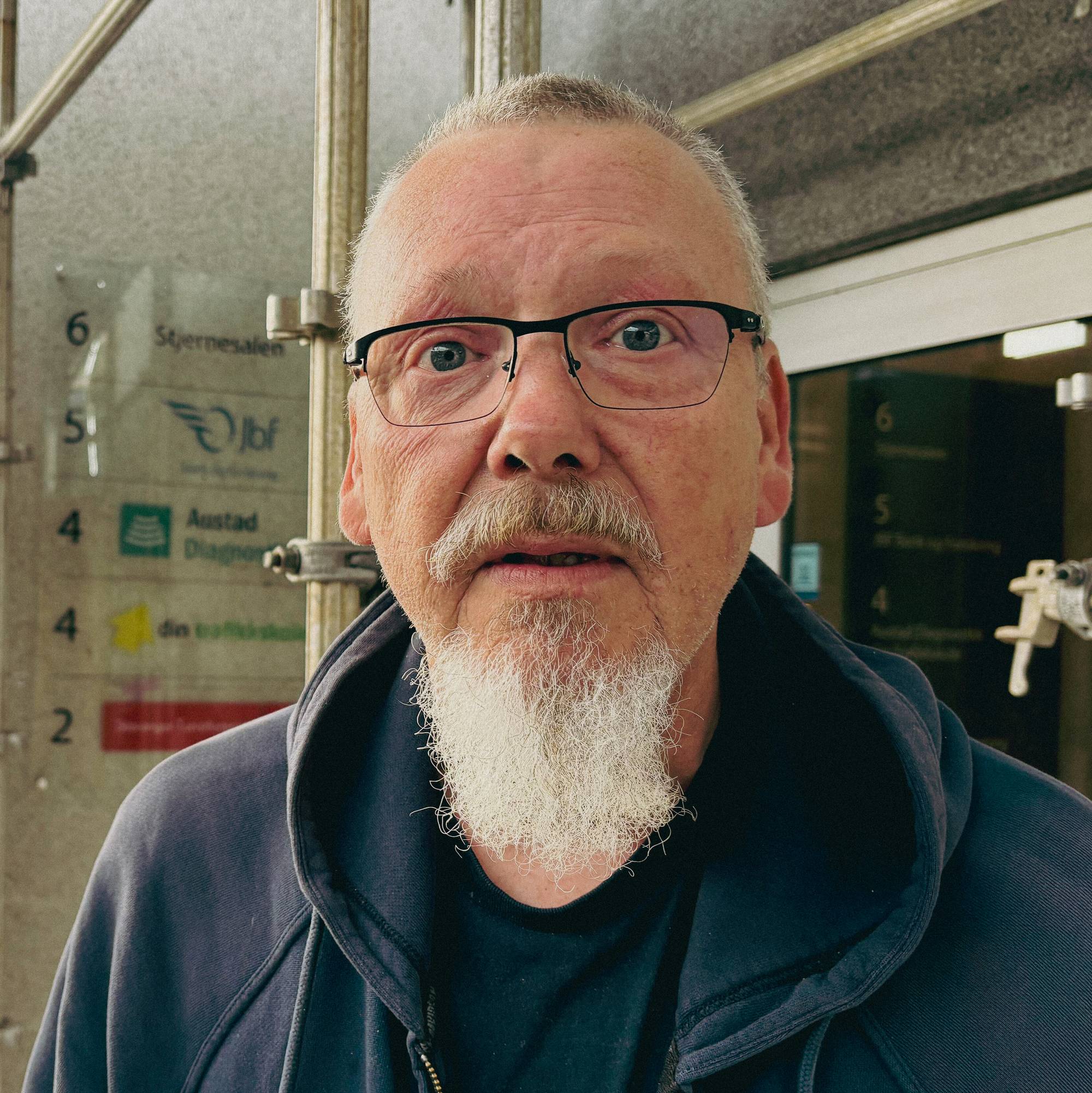 Mann med briller og skjegg står utenfor en bygning. Bakgrunnen viser en inngang og et bymiljø.