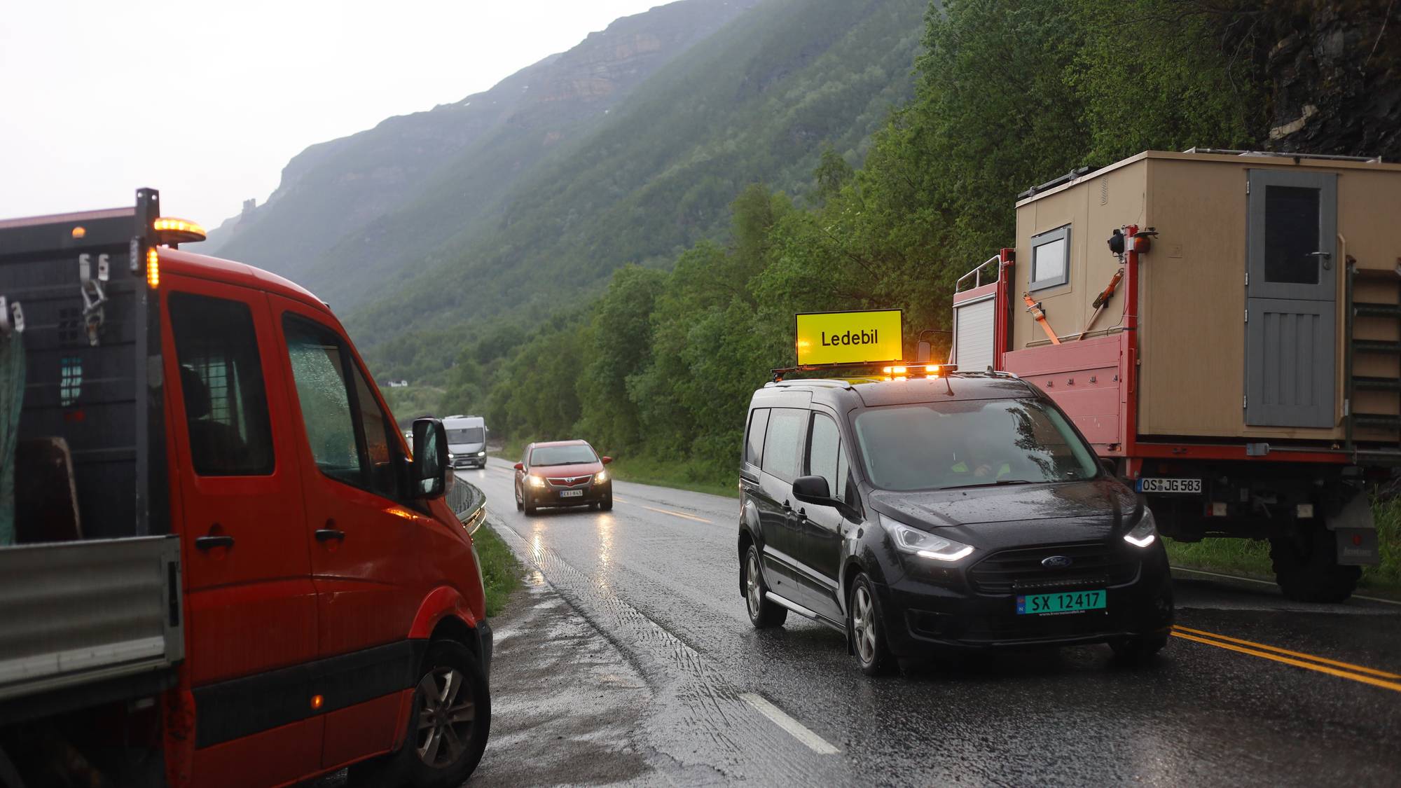 En ledebil med skiltet "Ledebil" kjører på en våt vei. Bak ledebilen står en lastebil med et påbygg som ligner et lite hus. I forgrunnen er en rød kassebil med varsellys. Det er et fjellandskap i bakgrunnen. (Bildebeskrivelsen er laget av en KI-tjeneste)