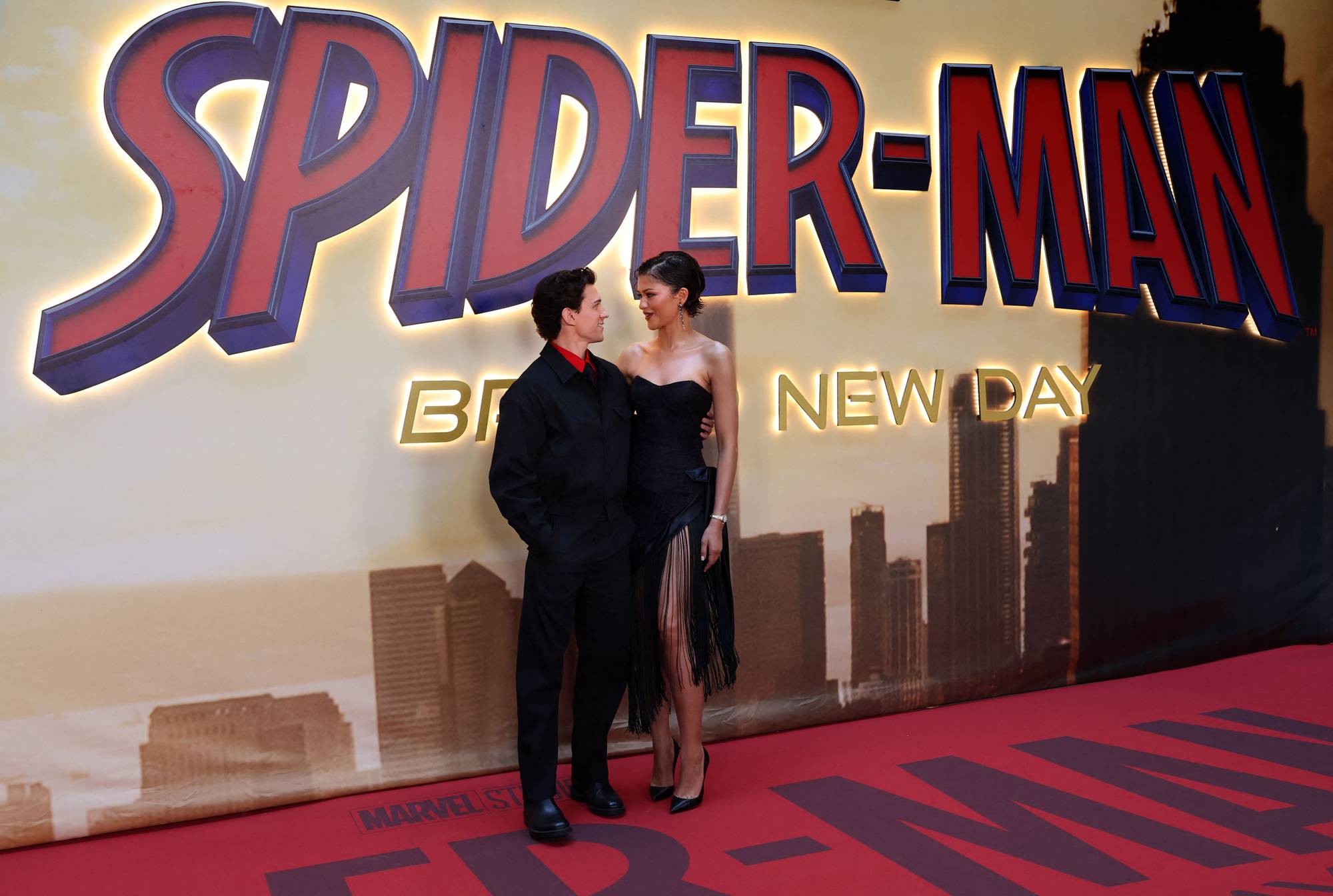 Skuespillerne Tom Holland og Zendaya poserer foran en stor "Spider-Man" logo. De er kledd i festantrekk. Bak dem er det en bakgrunn med en silhuett av en by og flere bygninger. Gulvet er rødt med "Spider-Man" logo gjentatt. (Bildebeskrivelsen er laget av en KI-tjeneste)