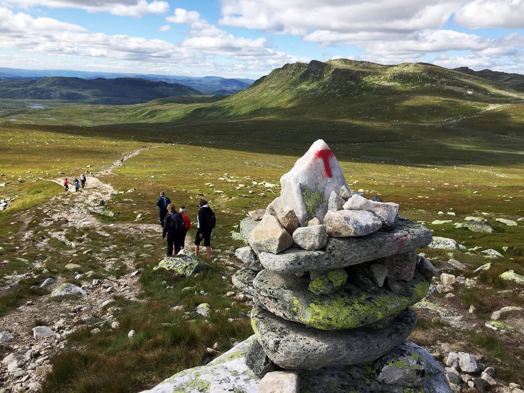 Turgåere på en fjellsti merket med en varde med rød T, med fjellandskap i bakgrunnen. (Bildebeskrivelsen er laget av en KI-tjeneste)