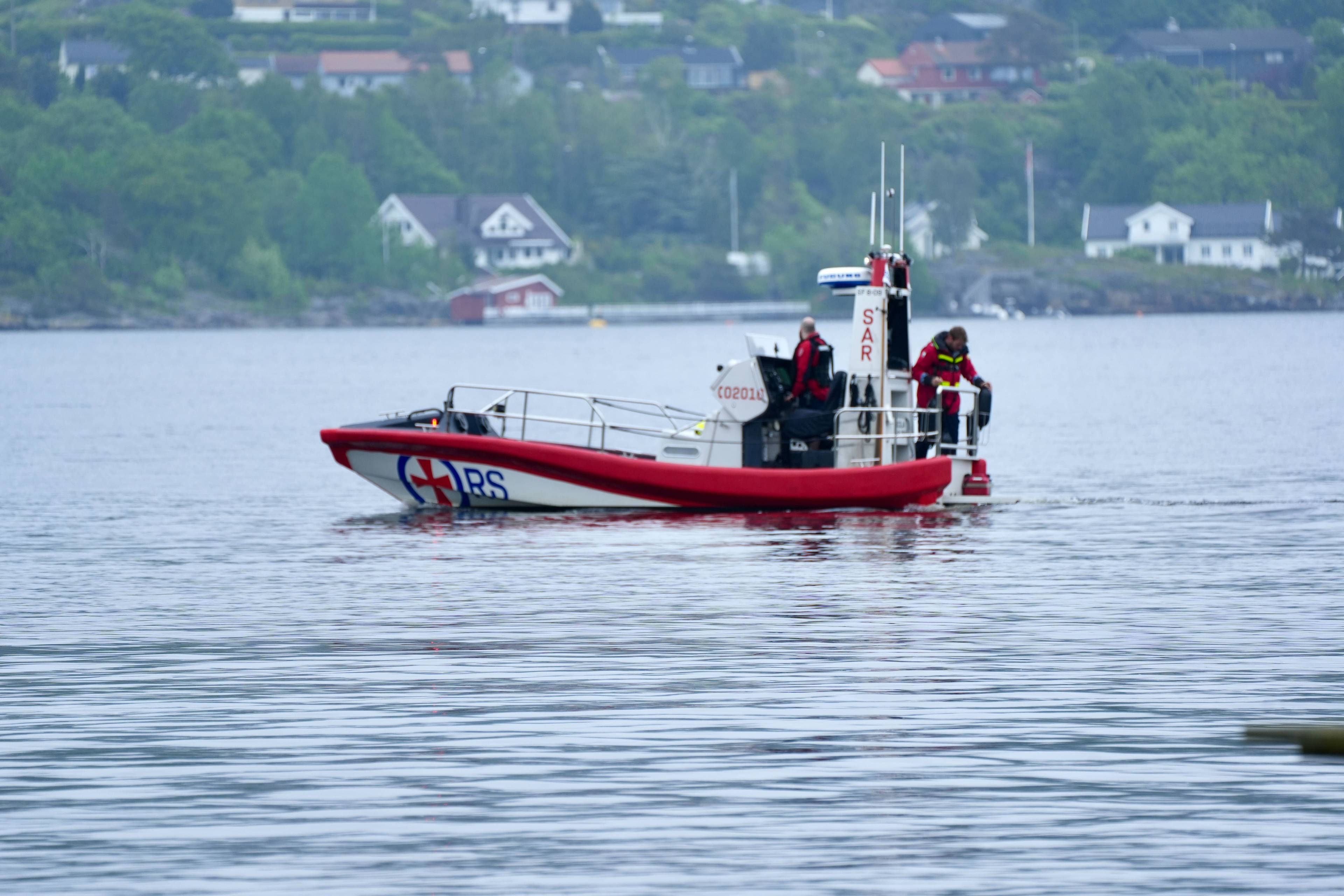 En redningsskøyte med to personer ombord er i fart på vannet. De er kledd i redningsvester og røde jakker. I bakgrunnen skimtes bebyggelse og trær langs en fjellside. (Bildebeskrivelsen er laget av en KI-tjeneste)