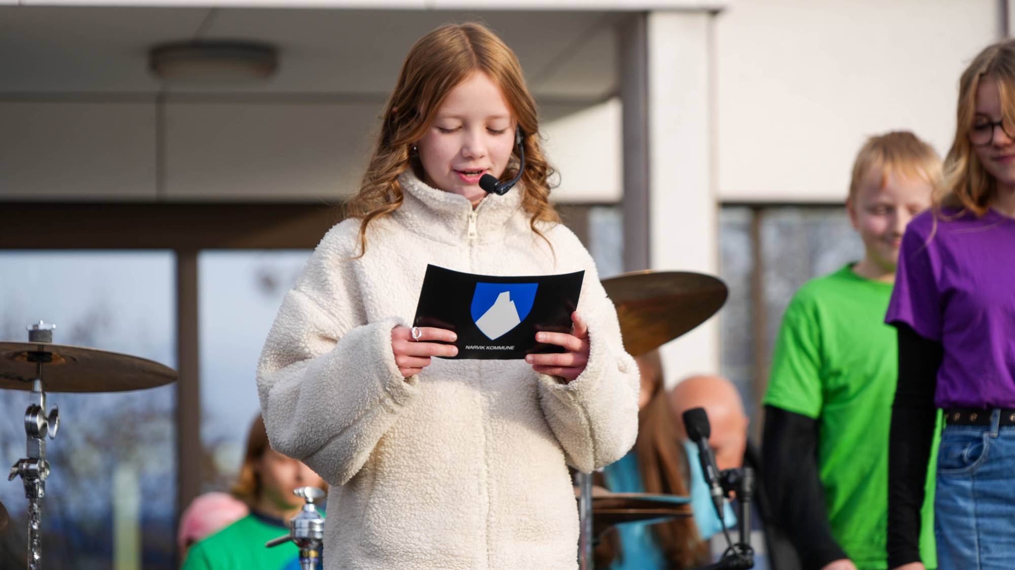 Mathilde Antonsen Myrvang (10) holder et korte i hendene og leser fra dette. I bakgrunn står flere barn og man ser deler av et trommesett.