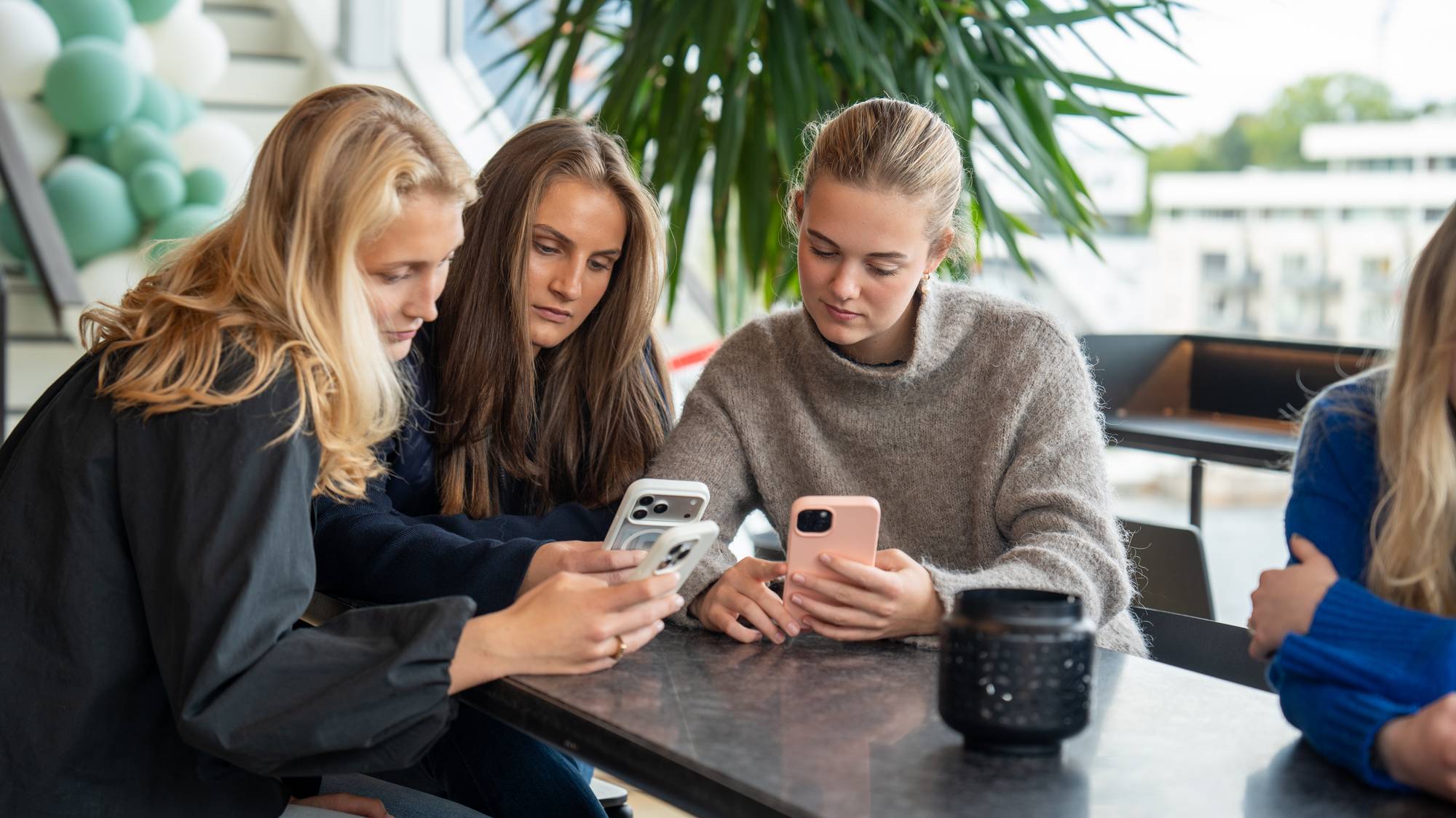 Tre unge kvinner sitter ved et bord og ser ned på hver sin mobiltelefon. (Bildebeskrivelsen er laget av en KI-tjeneste)