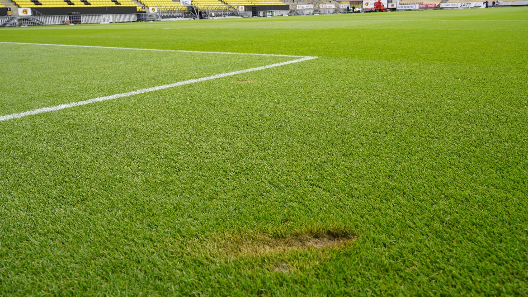 En tom fotballbane med gule tribuner i bakgrunnen. Et tydelig slitasjemerke er synlig i gresset. (Bildebeskrivelsen er laget av en KI-tjeneste)