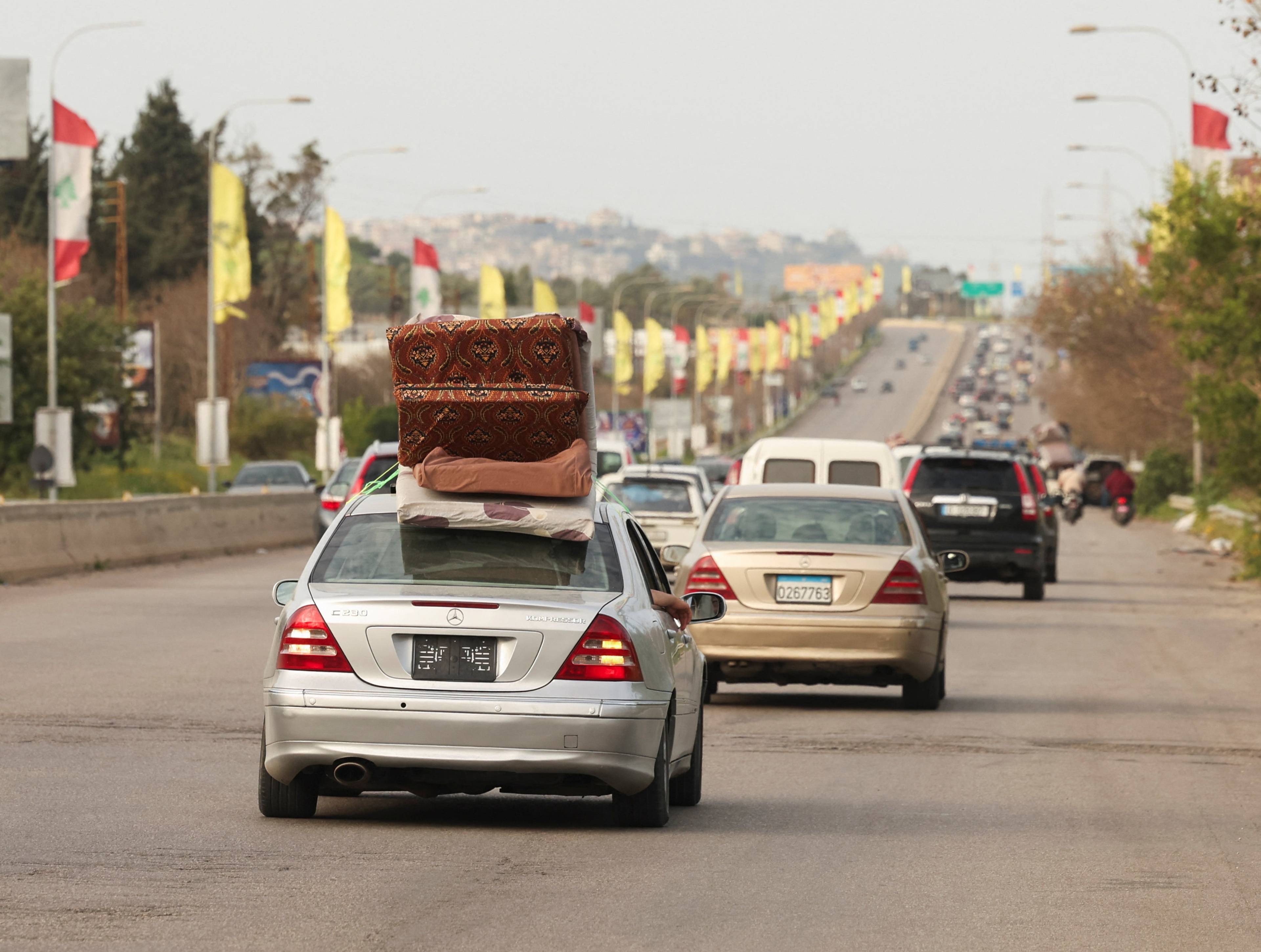 Biler med flyttelass på taket står i kø. En sølvfarget Mercedes-Benz er i forgrunnen. Langs veien er det flagg og lyktestolper. Trafikken flyter sakte fremover. (Bildebeskrivelsen er laget av en KI-tjeneste)