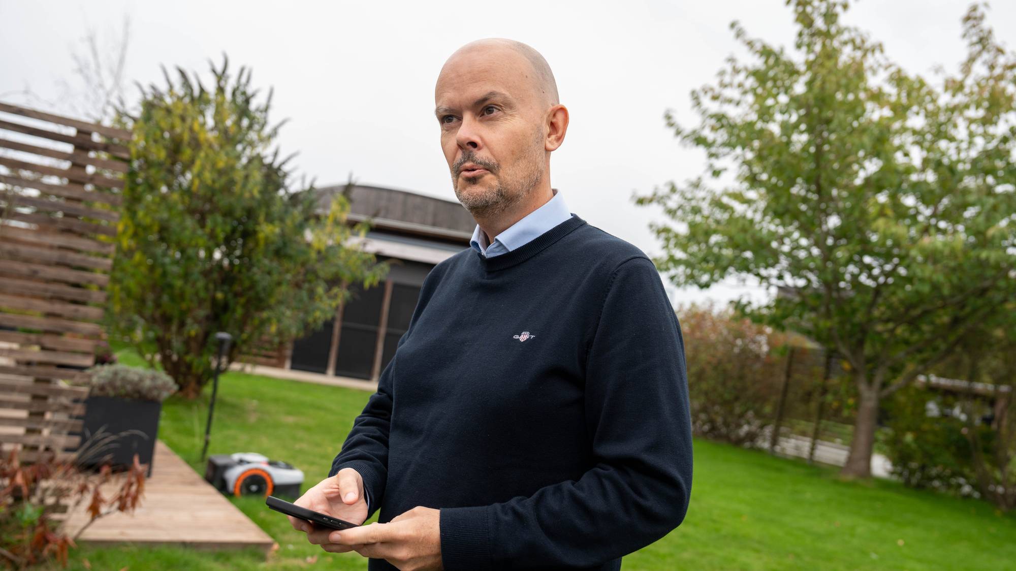 Mann med mobiltelefon i en hage der en robotgressklipper arbeider i bakgrunnen. (Bildebeskrivelsen er laget av en KI-tjeneste)