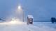 Mary Ann Holm Et busstopp ved en vei, ved en snødekt eng og snø i luften. (Bildebeskrivelsen er laget av en KI-tjeneste)