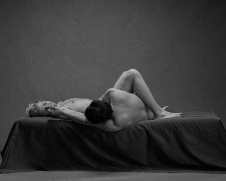 En naken mann med lyst hår ligger på ryggen i senga med knærne opp. En naken mann med mørkt hår ligger ved siden av han med kroppen under den lyshåredes knær.