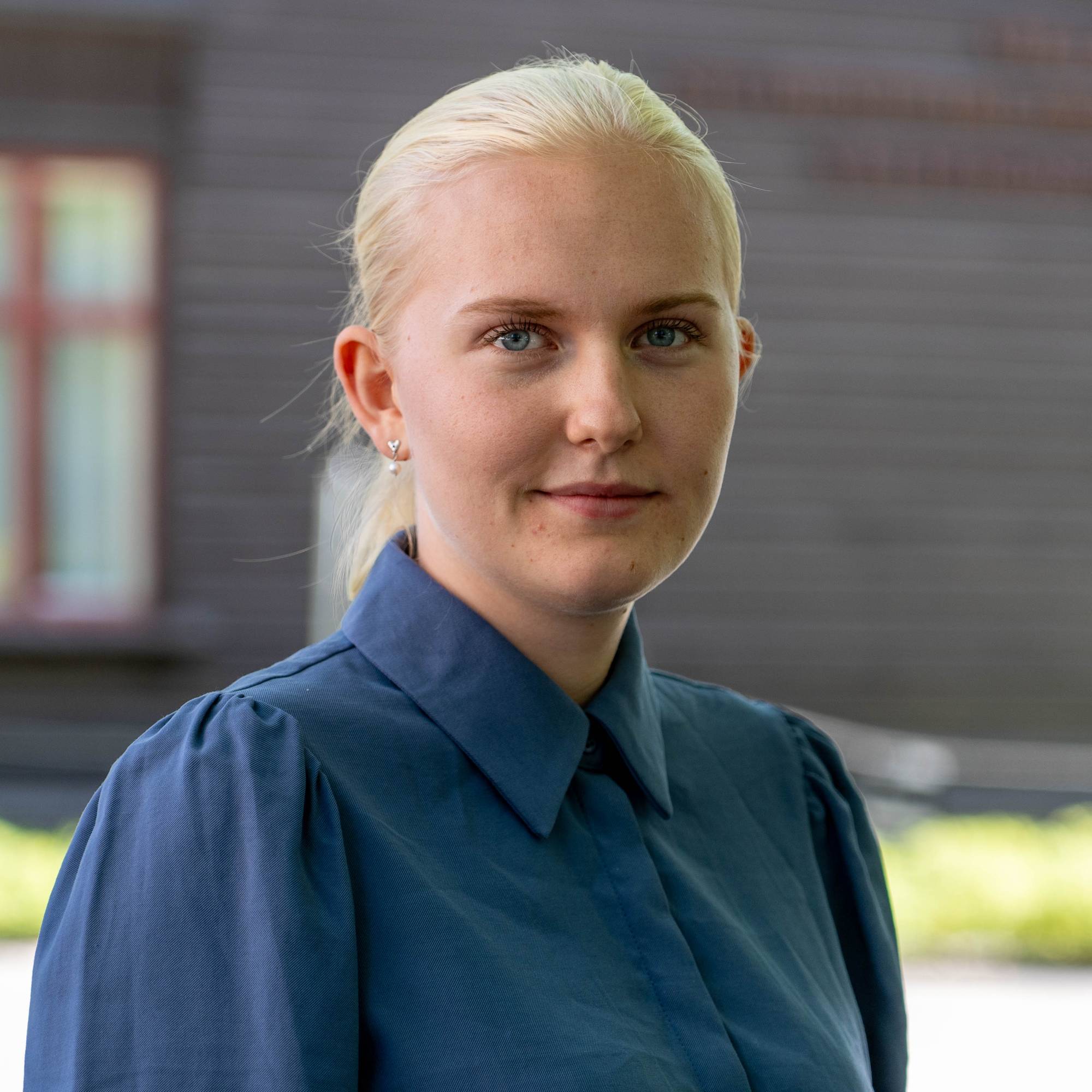 Portrett av en lyshåret kvinne i blå skjorte, med en mørk bygning i bakgrunnen. (Bildebeskrivelsen er laget av en KI-tjeneste)