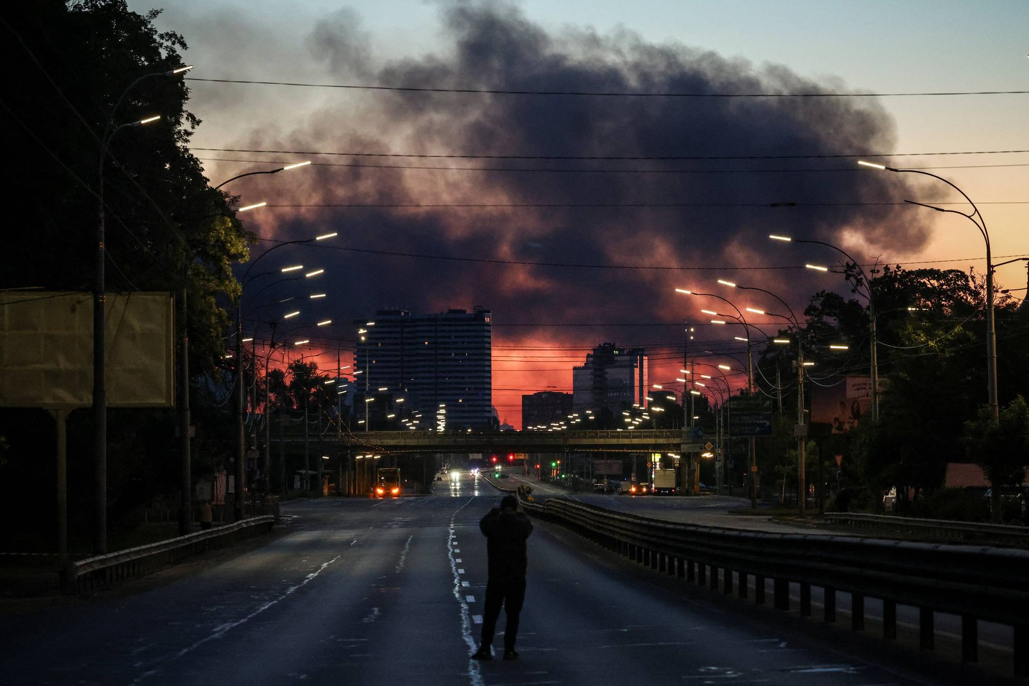 En mann står på en tom vei og fotograferer en stor røyksøyle som stiger mot en oransje himmel. Bakgrunnen viser bysilhuetter og en bro. (Bildebeskrivelsen er laget av en KI-tjeneste)